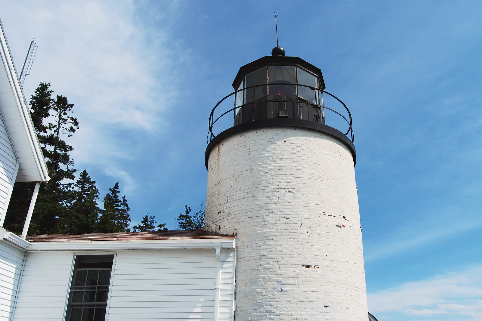 Der weiße Turm des Bass Harbor Head Light mit schwarzer Laterne vor blauem Himmel, davor das Wärterhaus