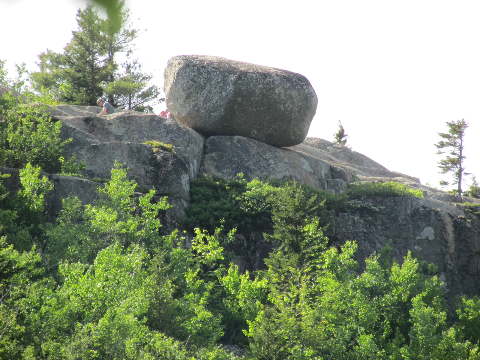 Der Findling Bubble Rock auf der Kante des South Bubble, mit Menschen daneben, über einer bewaldeten Felsflanke