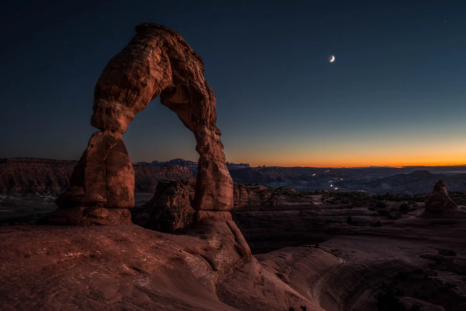 Der Delicate Arch in der Abenddämmerung vor tiefblauem Himmel mit schmaler Mondsichel