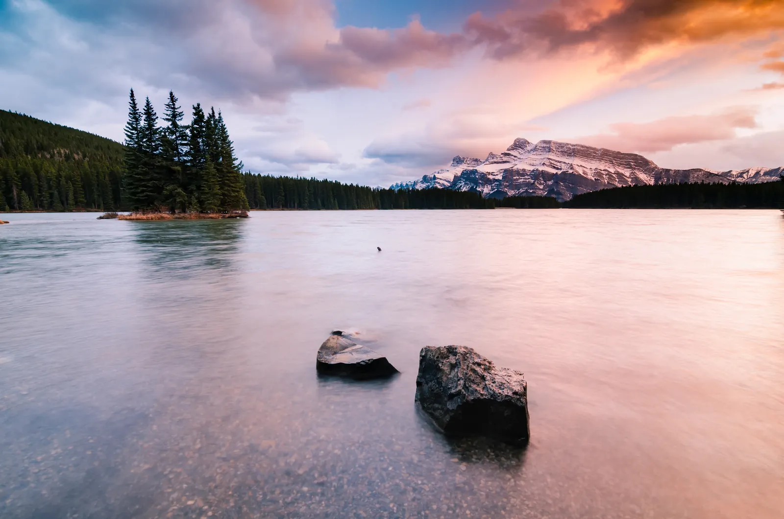 Two Jack Lake im Banff-Nationalpark im Abendlicht: rosa schimmerndes, glattes Wasser mit zwei Steinen, eine bewaldete Insel und verschneite Gipfel.