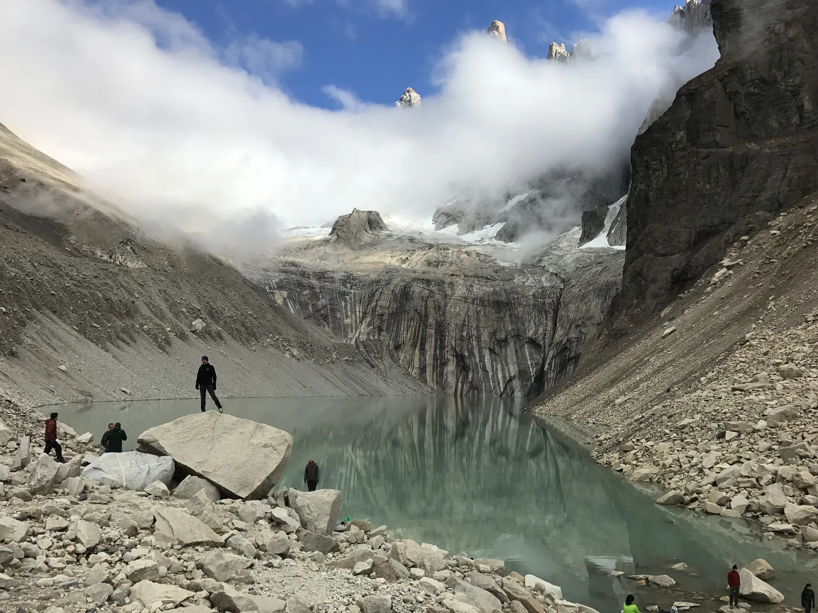 Wanderer am Mirador Base Torres im Nationalpark Torres del Paine: Felsblöcke am grünen Gletschersee, darüber Wolken an den Granittürmen