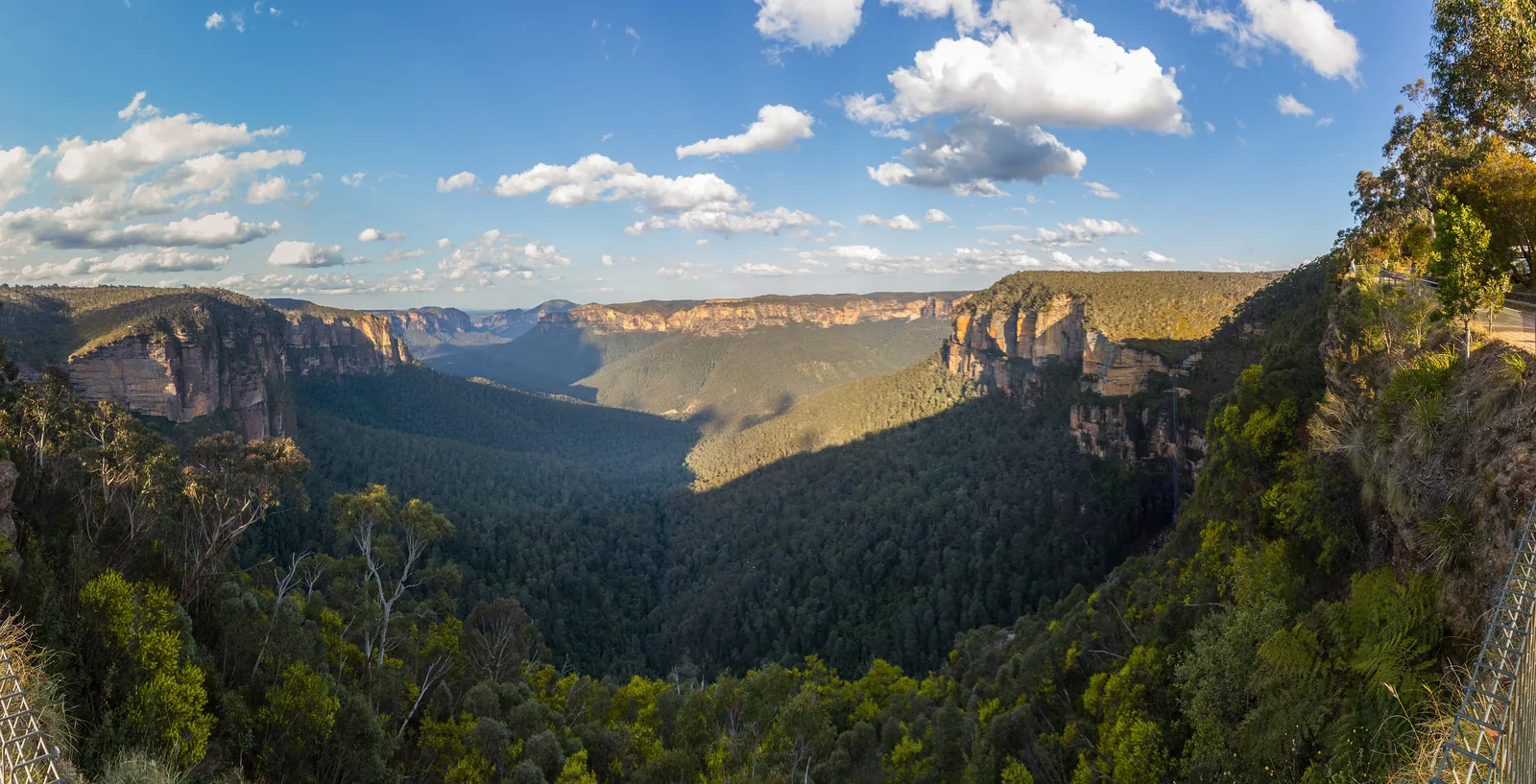 Panorama vom Govetts Leap im Blue-Mountains-Nationalpark: Sandsteinwände und das bewaldete Grose Valley im Abendlicht, rechts das Geländer der Aussicht