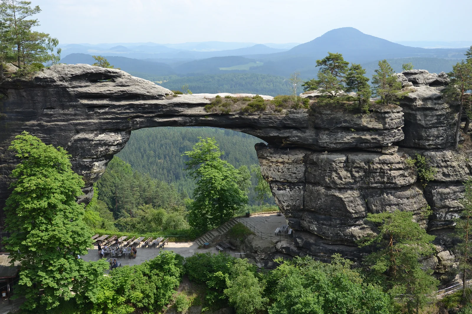Das Prebischtor im Nationalpark Böhmische Schweiz: ein mächtiger Sandsteinbogen über bewaldetem Grund, am Horizont der Kegel des Rosenbergs