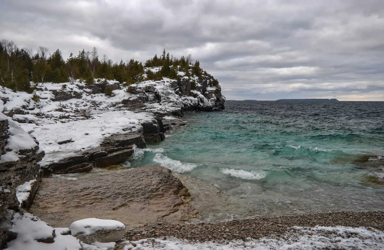 Verschneite Kalksteinterrassen am Indian Head Cove, darunter türkises Wasser, dahinter ein bewaldeter Felsvorsprung unter grauem Himmel