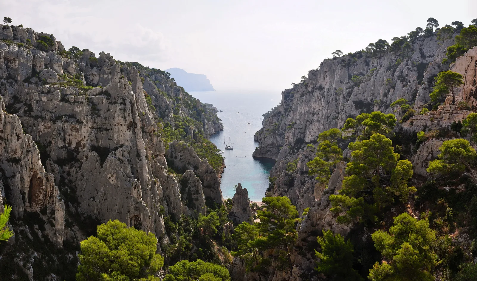 Die Calanque d’En-Vau im Nationalpark Calanques von oben: grüne Kiefern auf hellen Kalkwänden, unten die schmale Bucht mit Booten, hinten das Meer