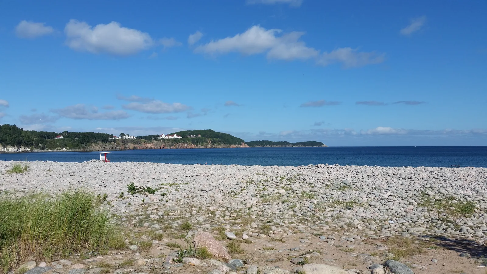 Breiter Kieselstrand am Freshwater Lake unter blauem Himmel, dahinter eine flache bewaldete Landzunge und das offene Meer