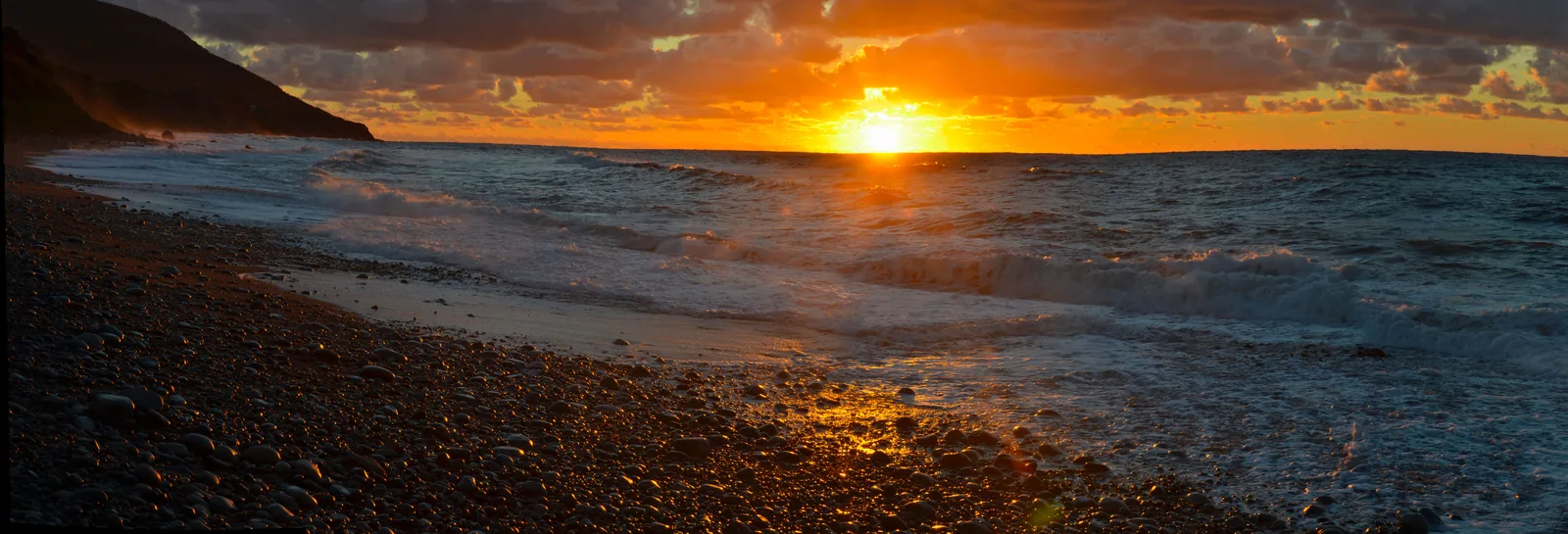 Sonnenuntergang über dem Wasser, gesehen von einem dunklen Kieselstrand; links läuft eine Steilküste ins Bild