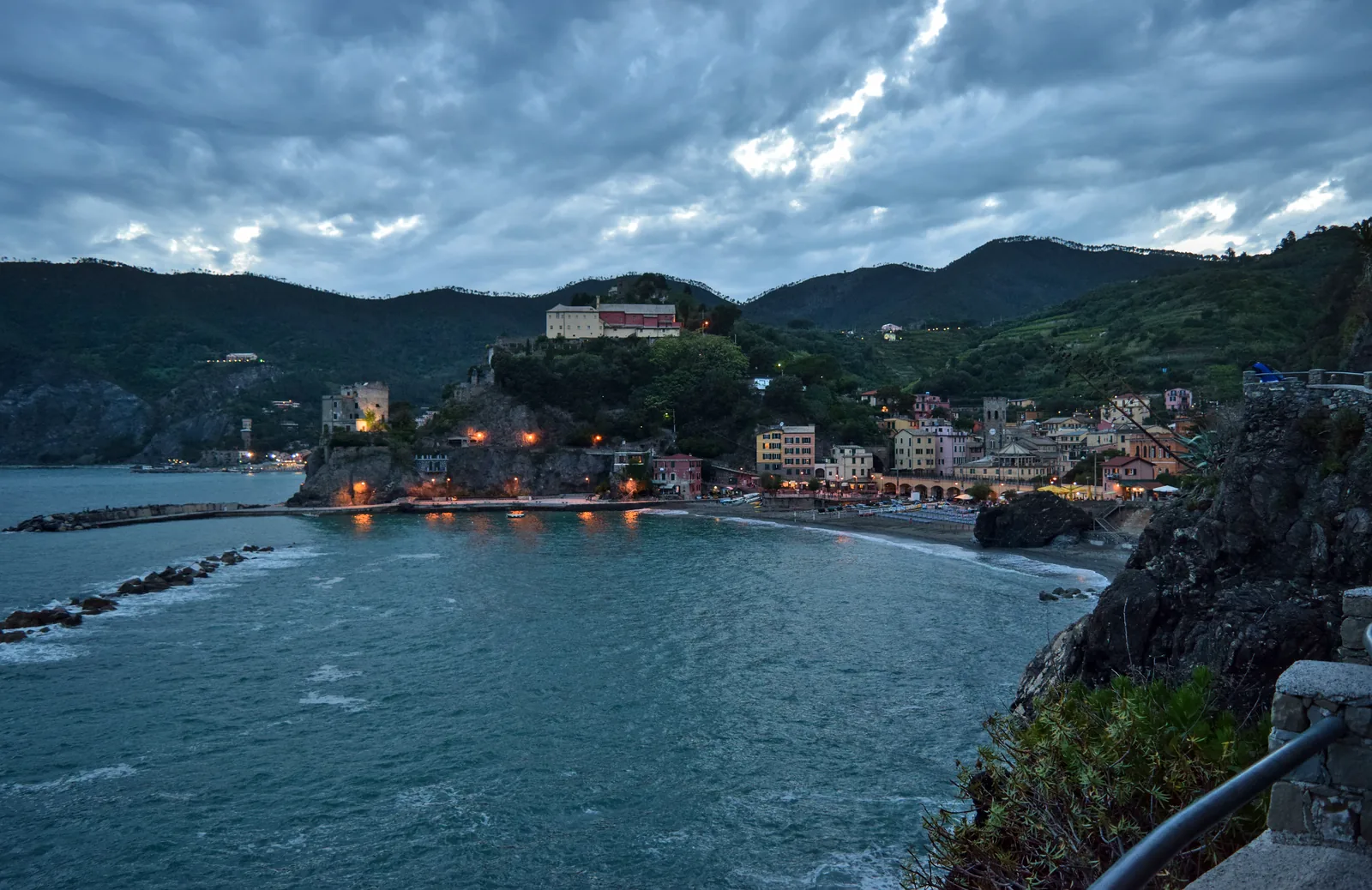 Abenddämmerung über dem Hafen von Monterosso al Mare im Nationalpark Cinque Terre, dahinter Lichter des Dorfs und bewaldete Hügel
