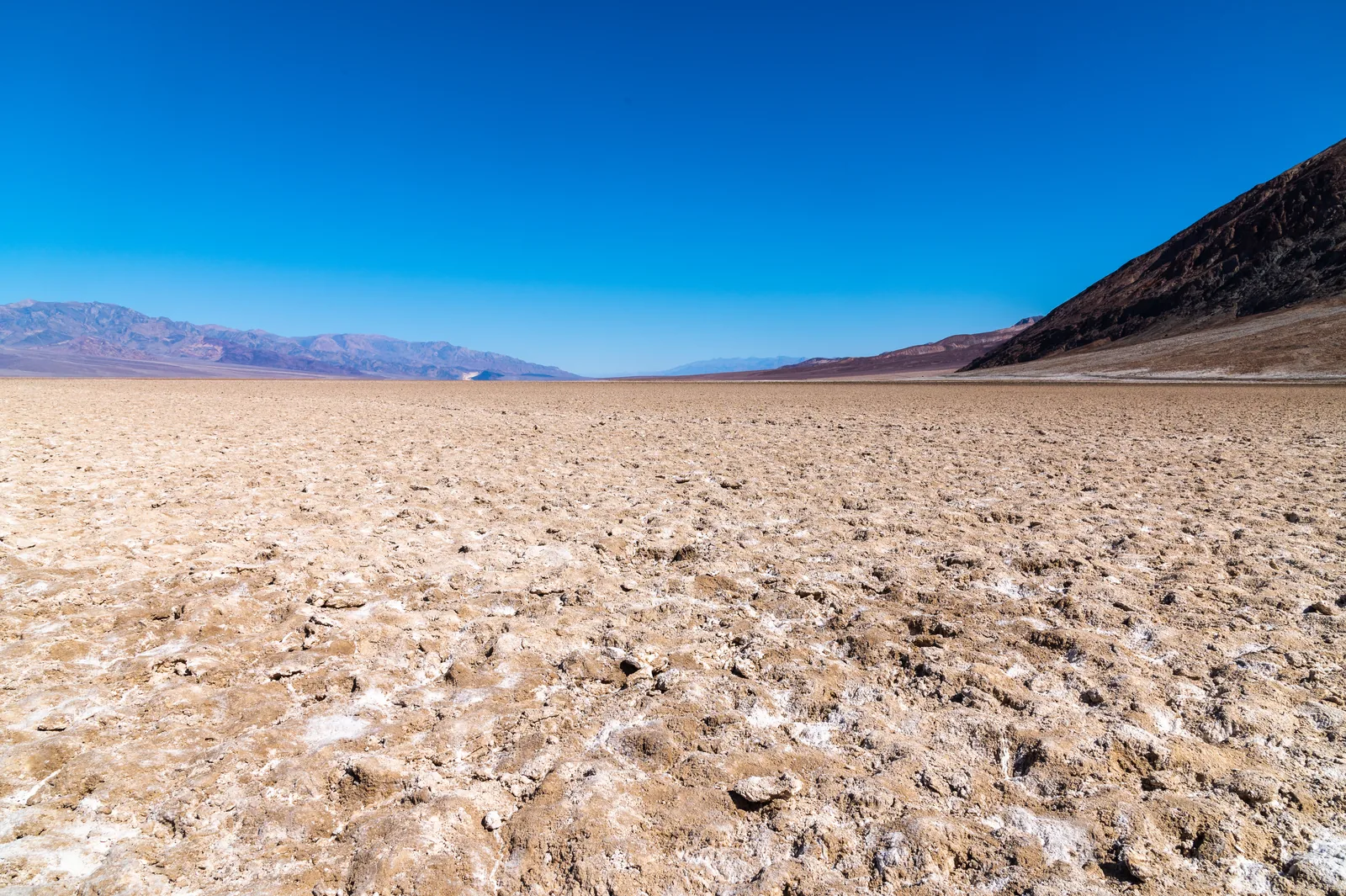 Aufgebrochene weiß-braune Salzkruste des Badwater Basin unter tiefblauem Himmel, rechts ein dunkler Berghang