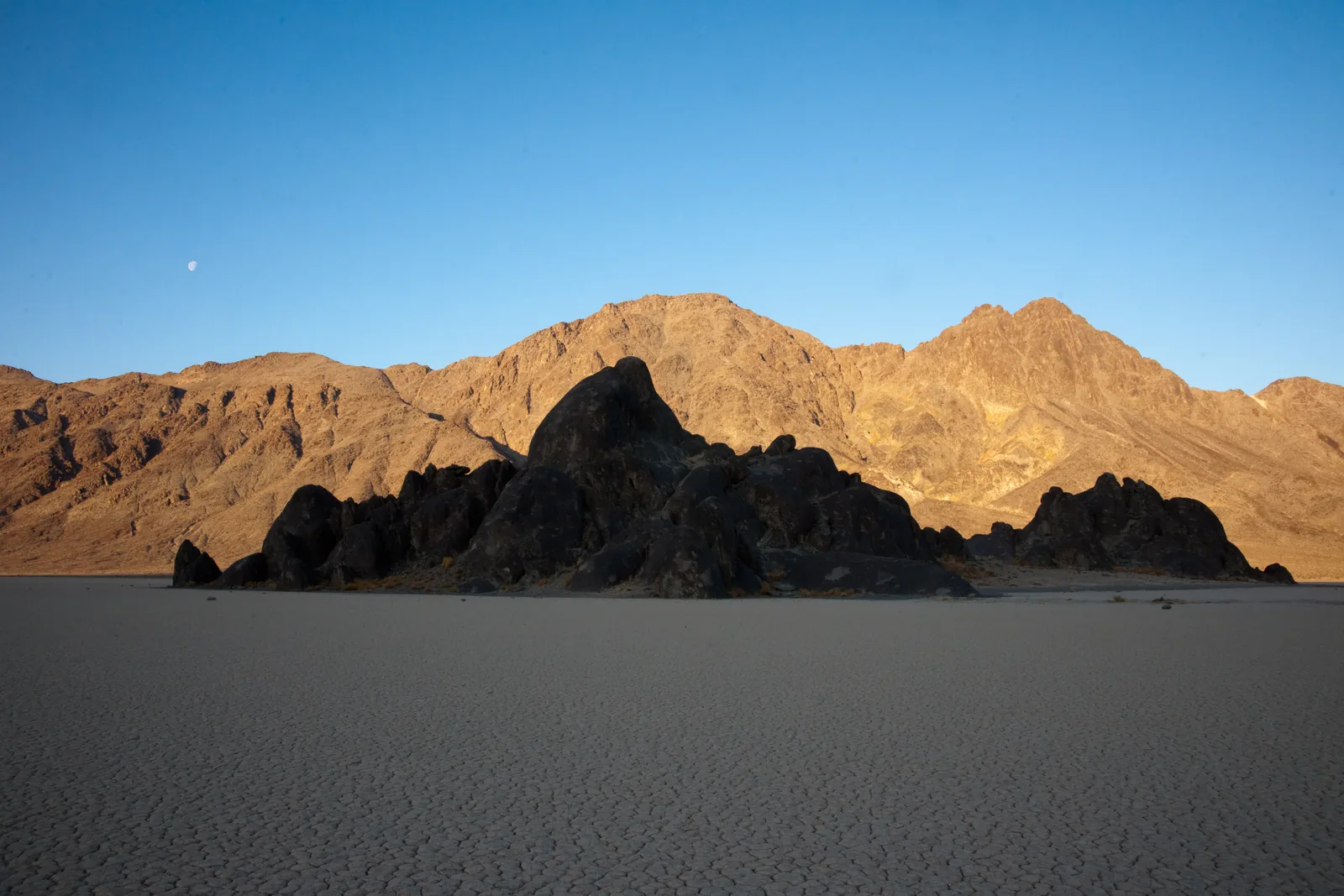 Die dunkle Felsinsel The Grandstand ragt aus der flachen grauen Racetrack Playa, dahinter von der Morgensonne beleuchtete Berge