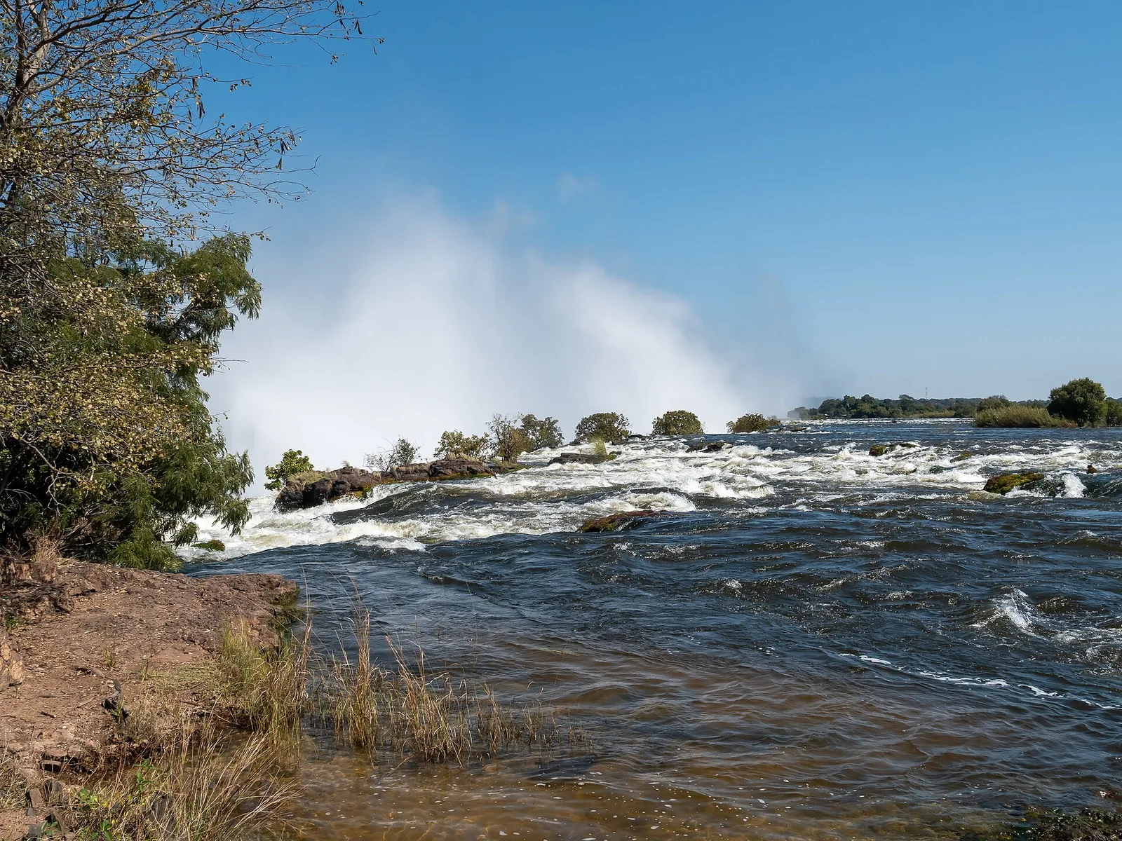 Der Sambesi fließt auf der sambischen Seite über Felsen zur Kante der Victoria Falls, dahinter steigt Gischt auf