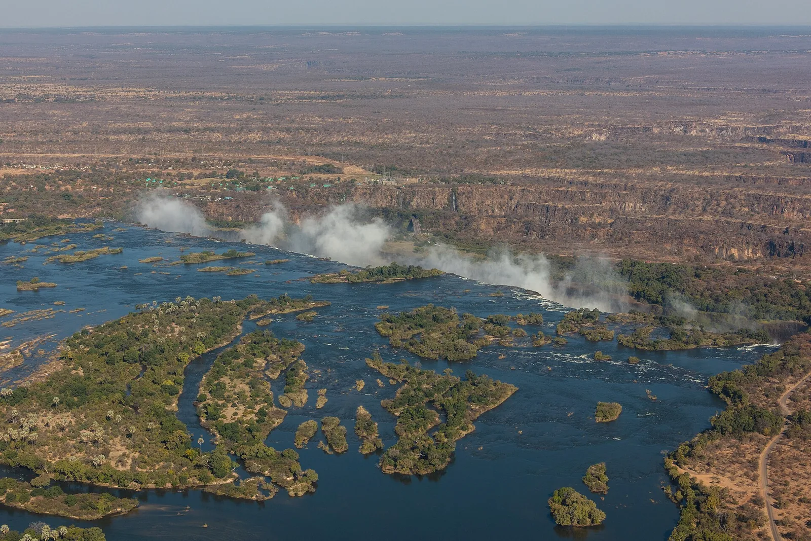 Luftbild des Sambesi oberhalb der Victoria Falls mit bewaldeten Inseln bis an die Kante, im Hintergrund Gischtwolken