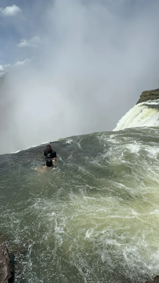 Ein Schwimmer im Devil's Pool an der Kante der Victoria Falls, vorn das grüne Wasser des Beckens, dahinter die Gischt