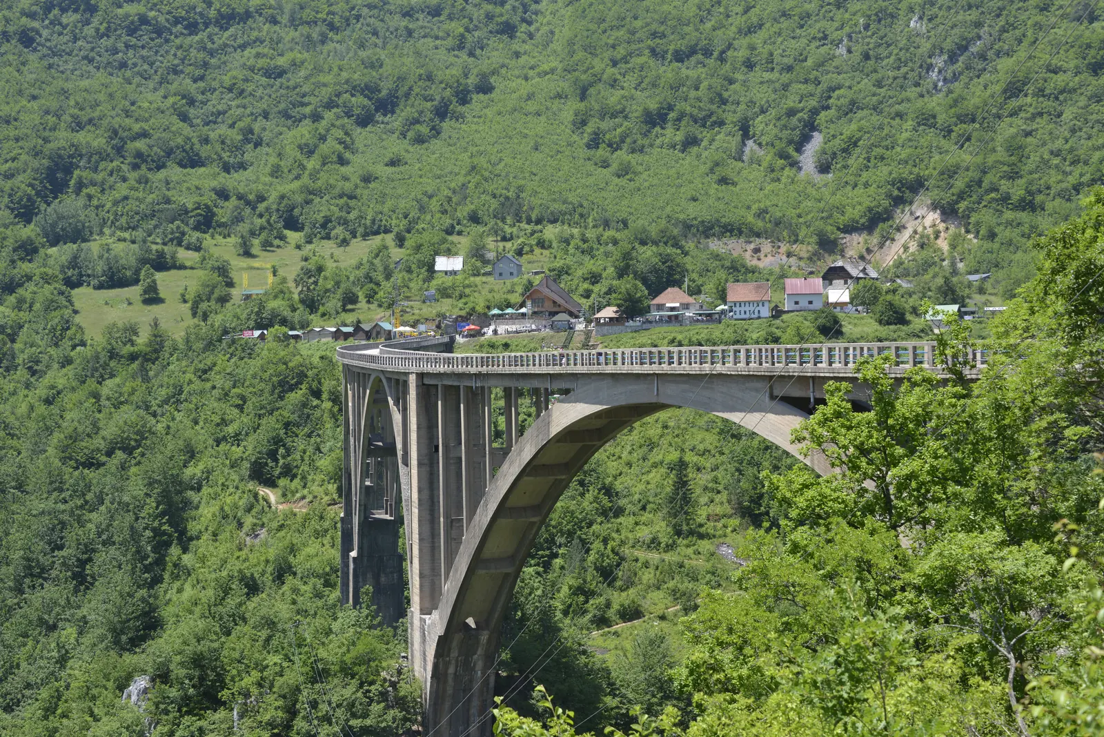 Eine hohe Betonbogenbrücke überspannt eine dicht bewaldete Schlucht, am Ende der Brücke einige Häuser