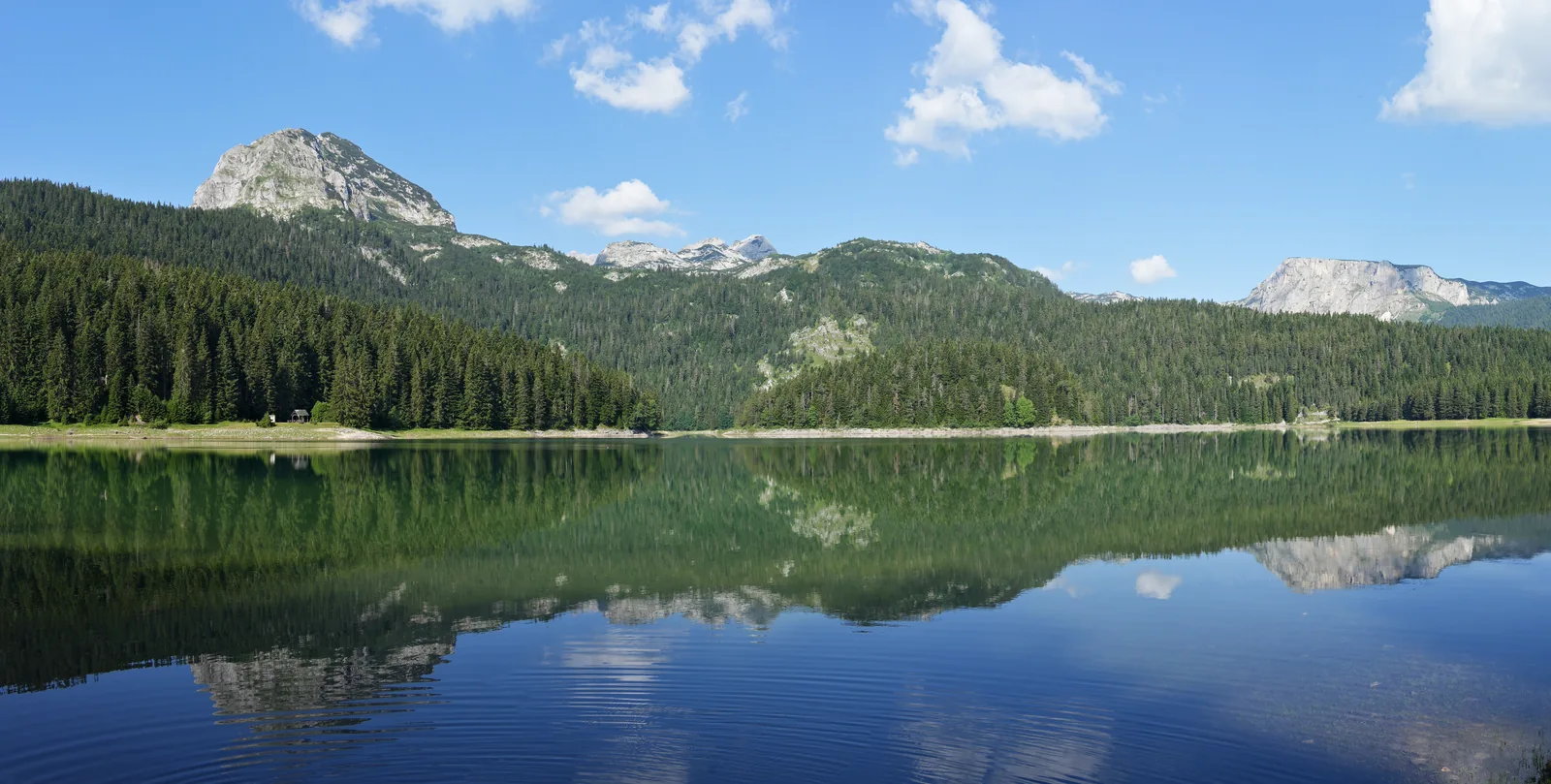 Der Schwarze See spiegelt glatt einen bewaldeten Uferhang und einen hellen Felsgipfel unter blauem Himmel mit Wolken