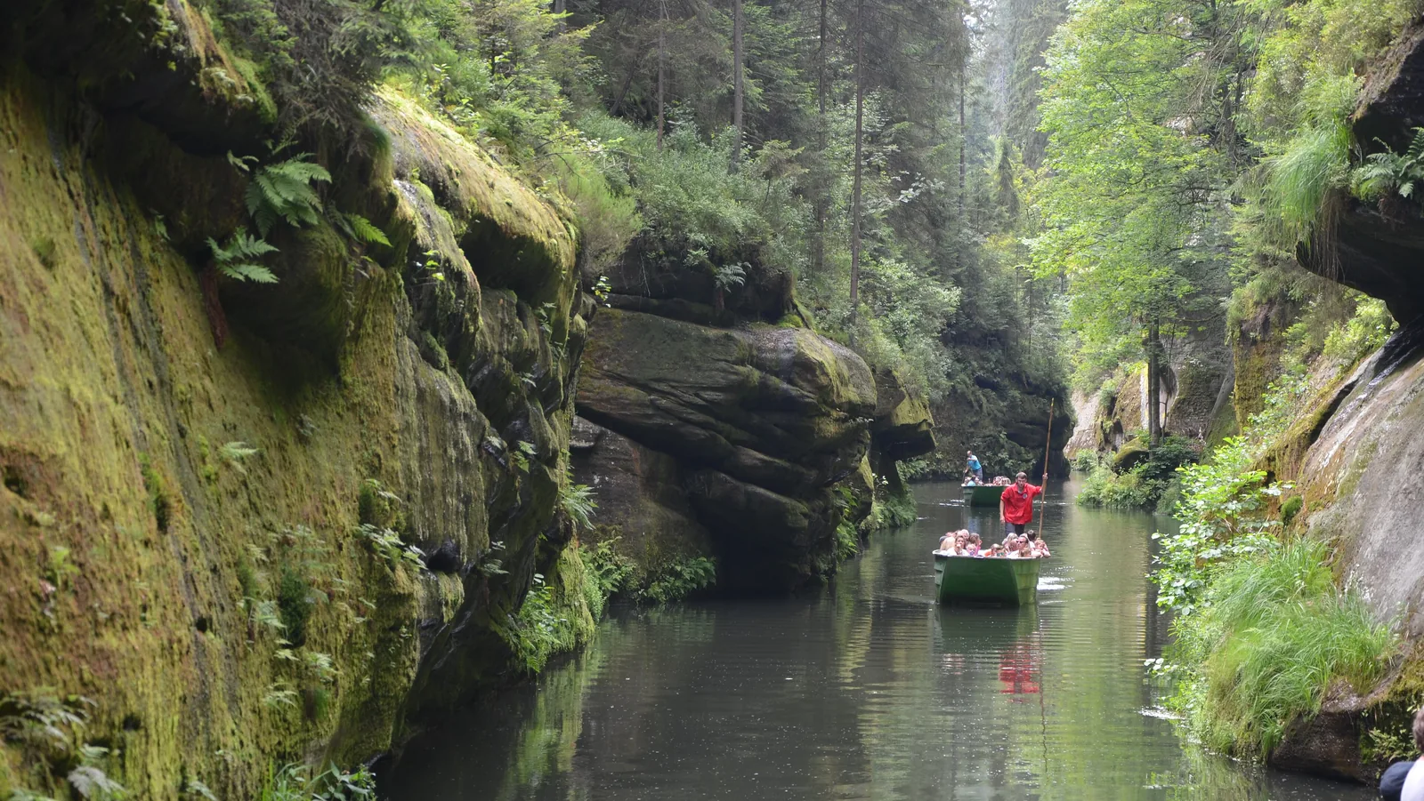Ein Kahn mit Fahrgästen und Fährmann in roter Jacke auf dem stillen Wasser der Edmundsklamm zwischen bemoosten Sandsteinwänden