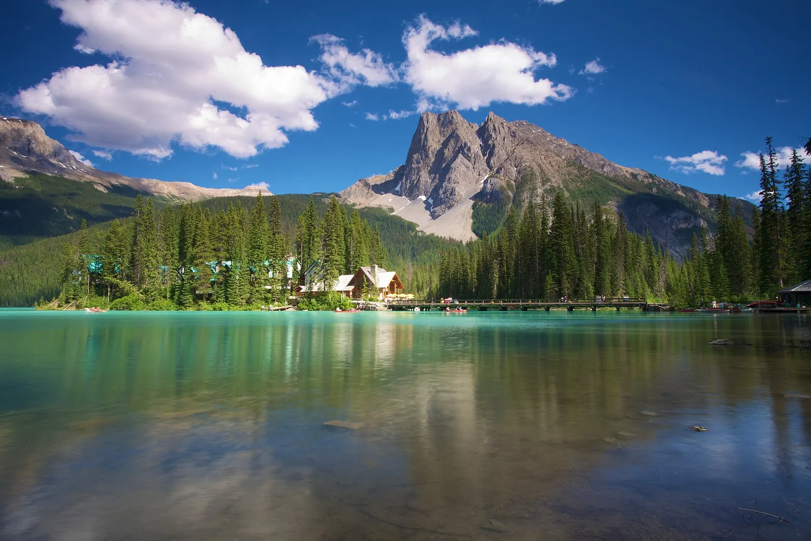 Emerald Lake im Yoho-Nationalpark mit der bewaldeten Halbinsel der Lodge vor einem Felsgipfel, im Vordergrund ruhiges Wasser.