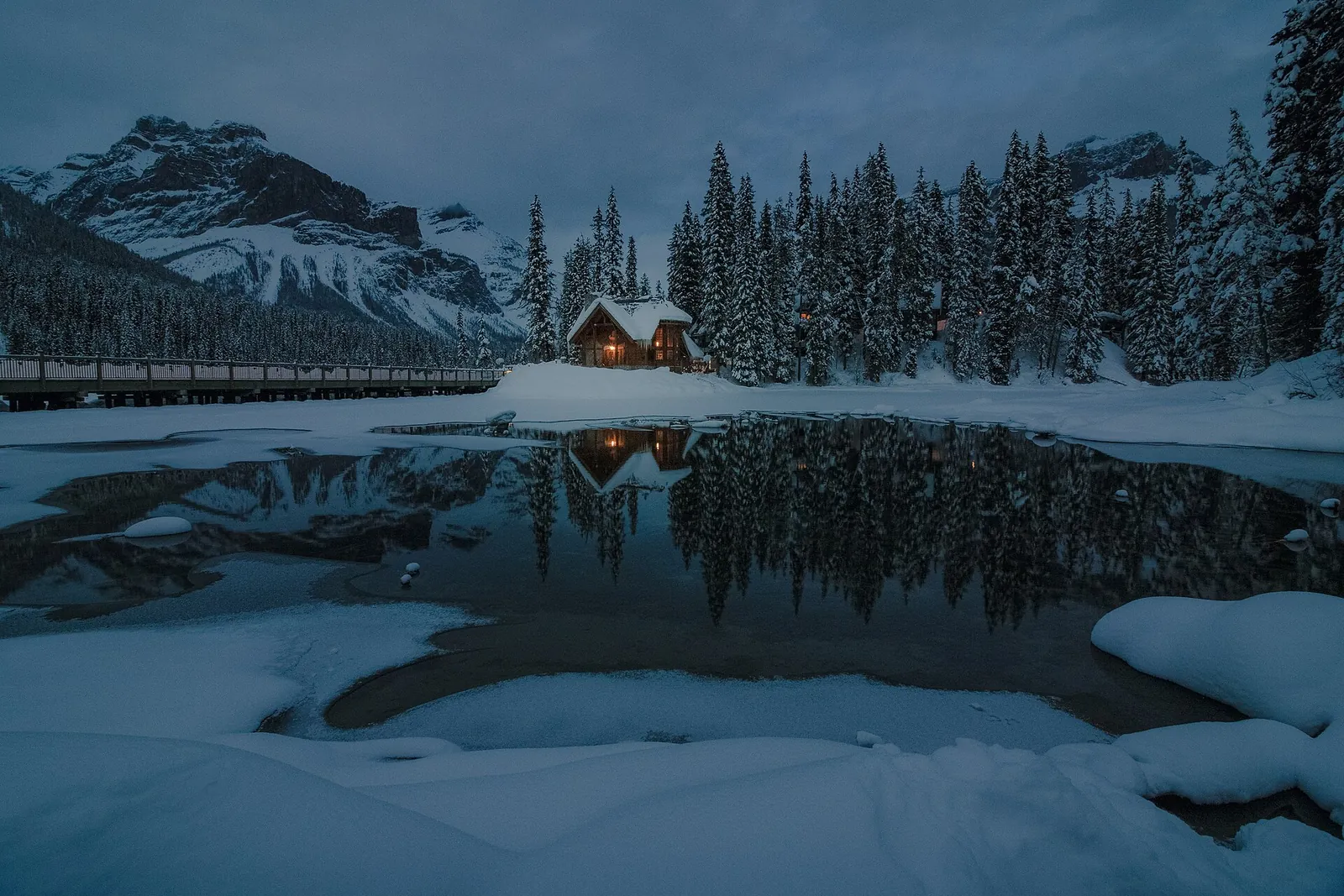 Der Emerald Lake im Yoho-Nationalpark im Winter: verschneites Ufer, offenes dunkles Wasser und eine beleuchtete Hütte zwischen Tannen.