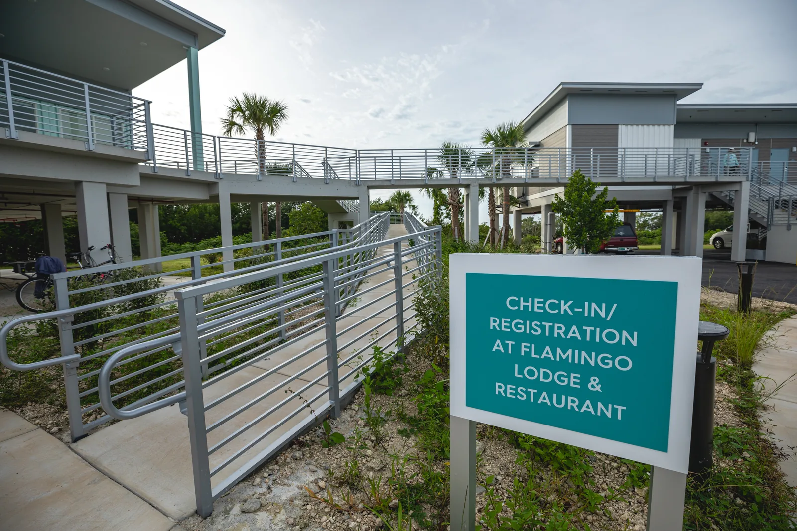 Neubauten der Flamingo Lodge auf Stelzen mit Rampen, davor ein türkises Schild für den Check-in von Lodge und Restaurant