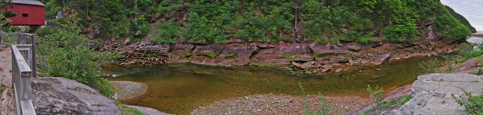 Breites Panorama des Point Wolfe River: rote Sandsteinwände, im Flussbett die Reste eines alten Staudamms, links am Bildrand die überdachte Brücke