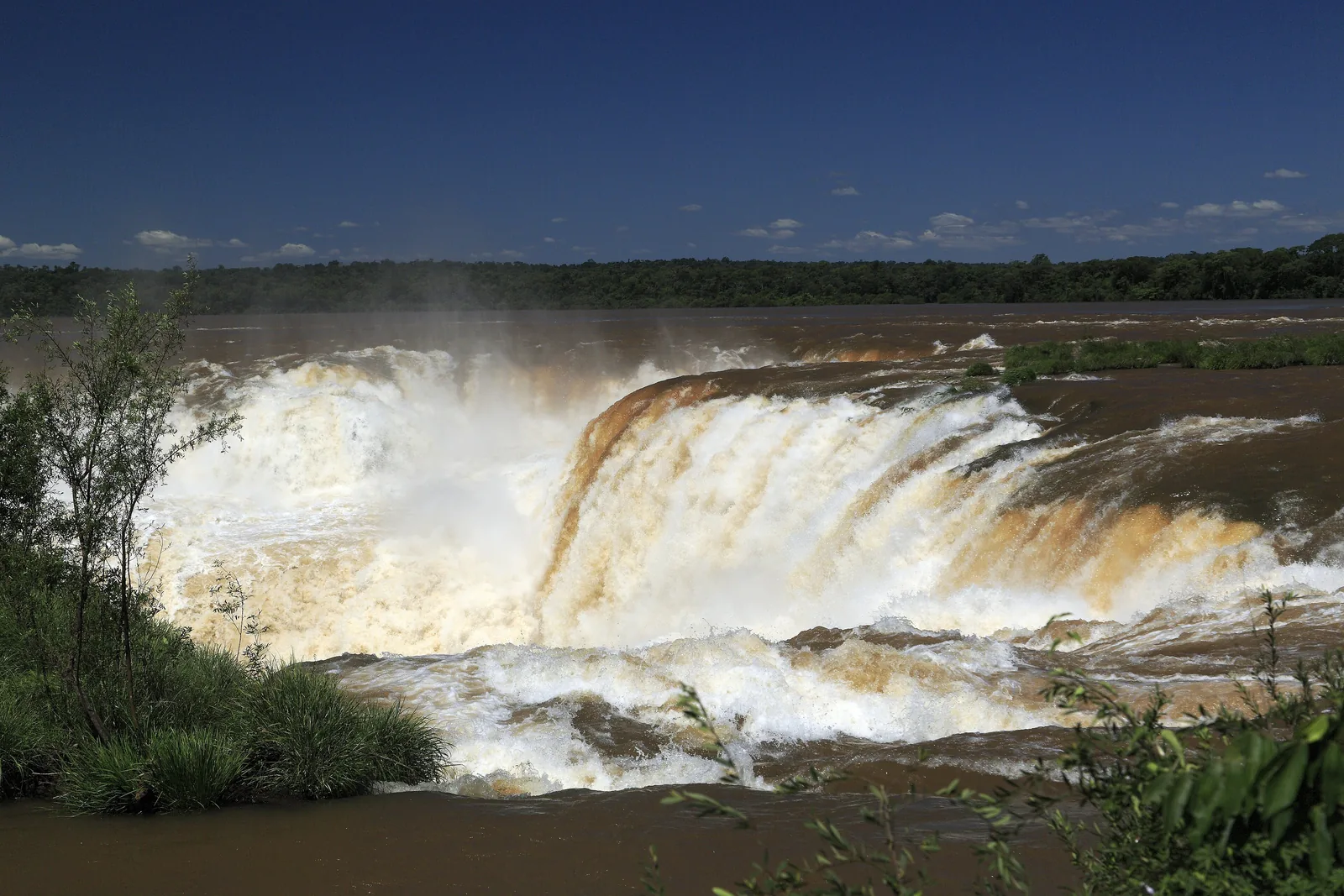Der Iguazú stürzt mit braunem Hochwasser in die Garganta del Diablo, Gischt steigt über die Kante, dahinter das argentinische Ufer