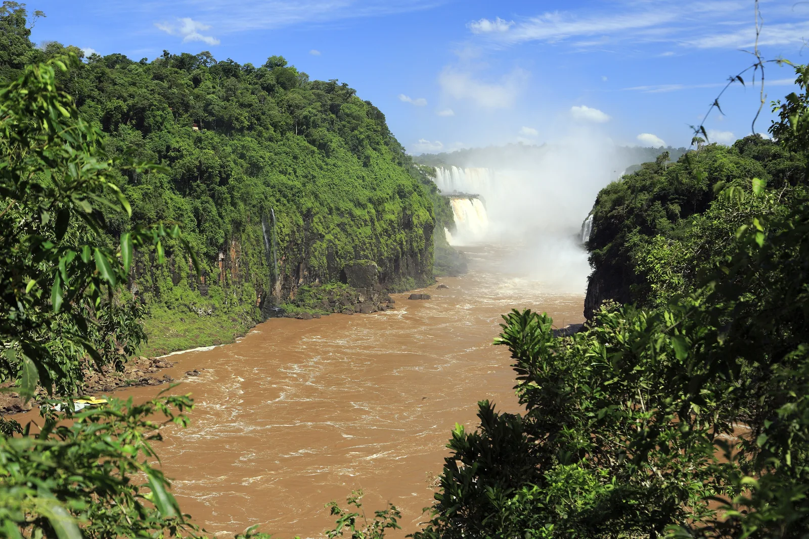 Der Iguazú fließt braun durch eine bewaldete Schlucht, im Hintergrund steigt Gischt aus der Garganta del Diablo
