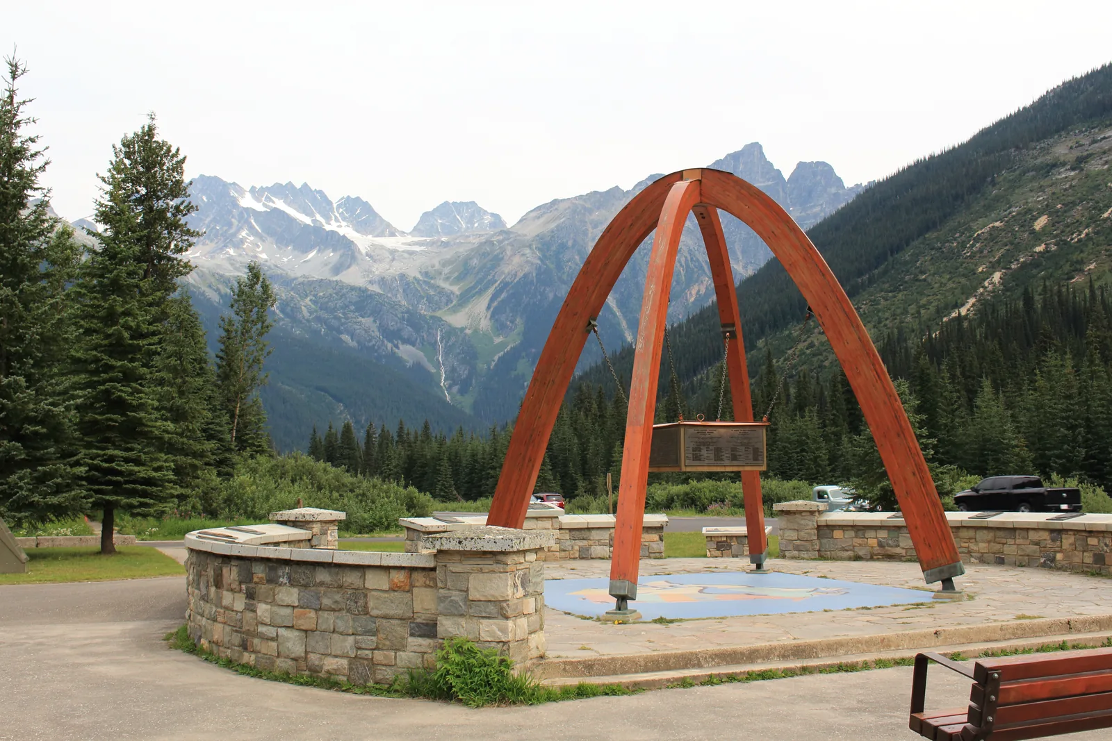 Hölzerner Bogen des Gipfeldenkmals am Rogers Pass, dahinter bewaldete Hänge und die Gletscher der Hermit-Kette