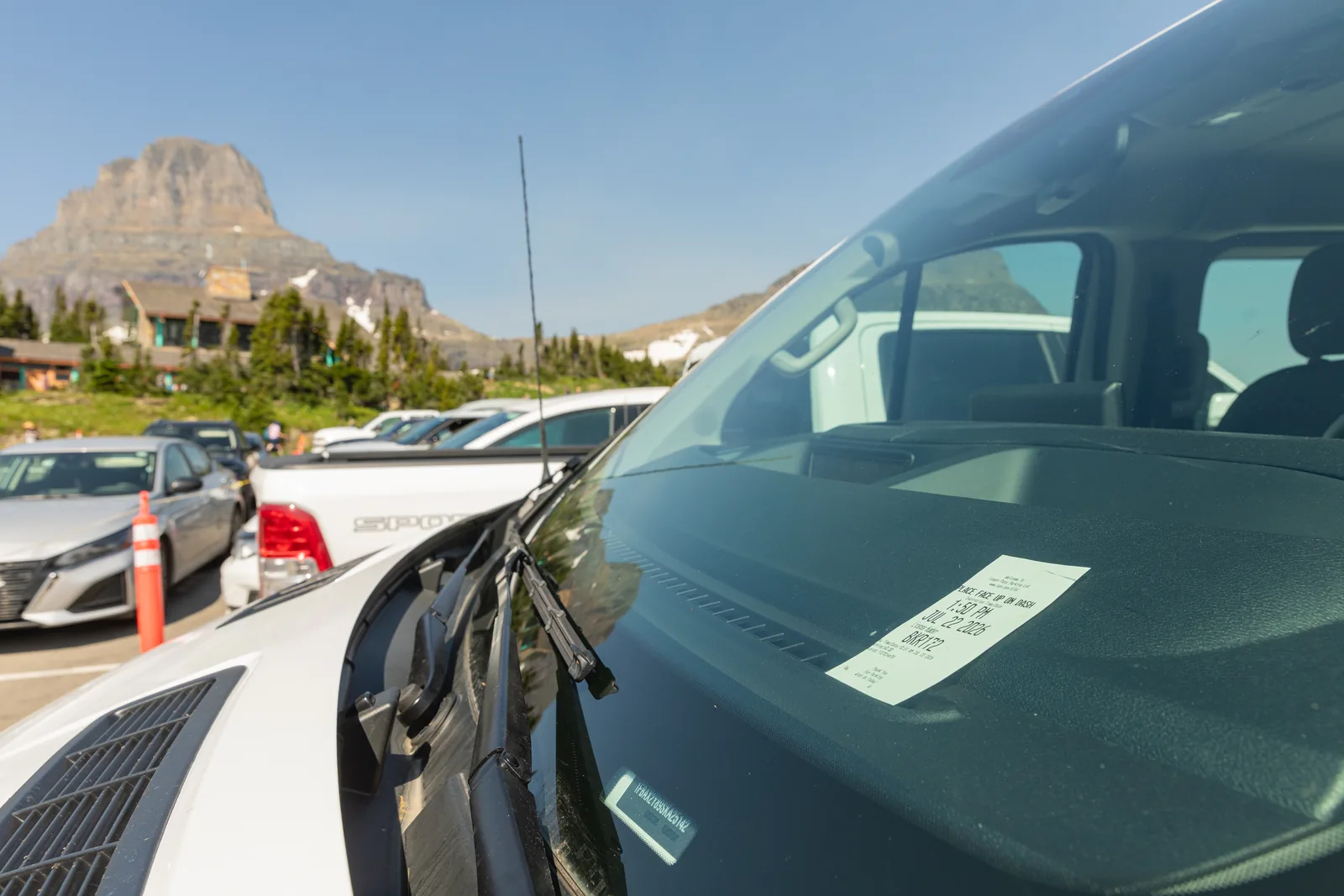 Ein Parkschein liegt hinter der Windschutzscheibe eines Autos auf dem vollen Parkplatz am Logan Pass, dahinter Besucherzentrum und Berge