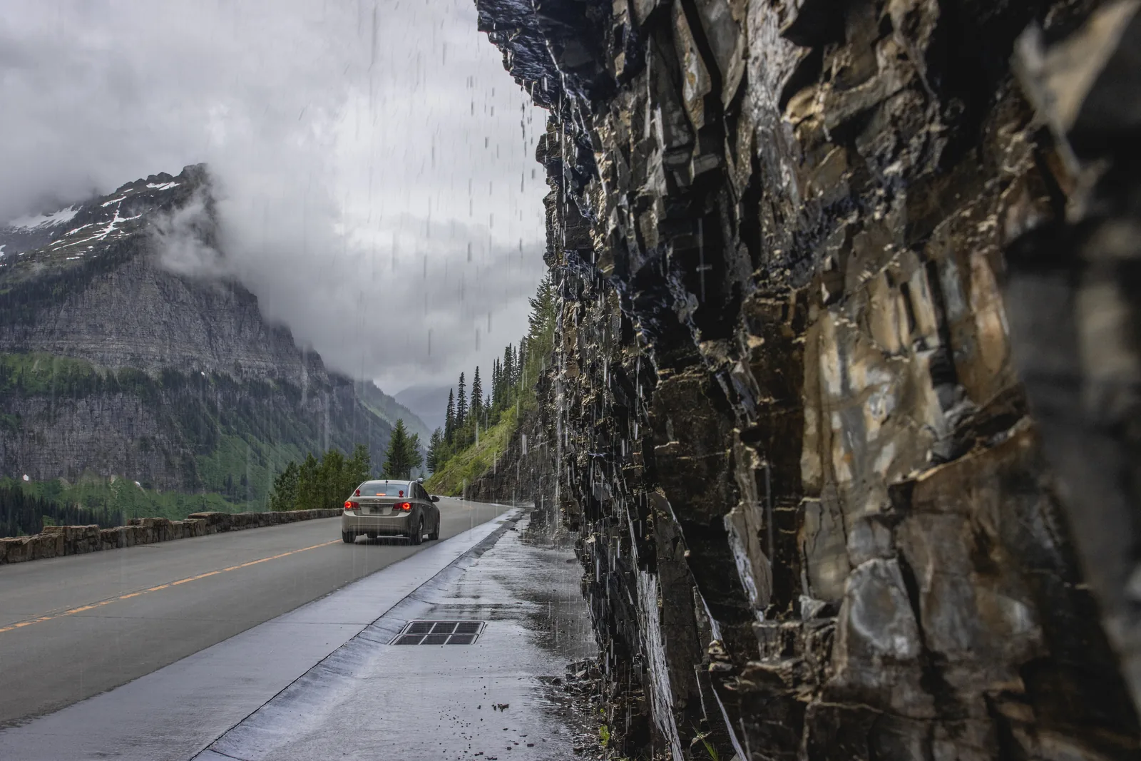 Wasser stürzt an der Weeping Wall direkt auf die nasse Fahrbahn der Going-to-the-Sun Road, ein Auto fährt vorbei