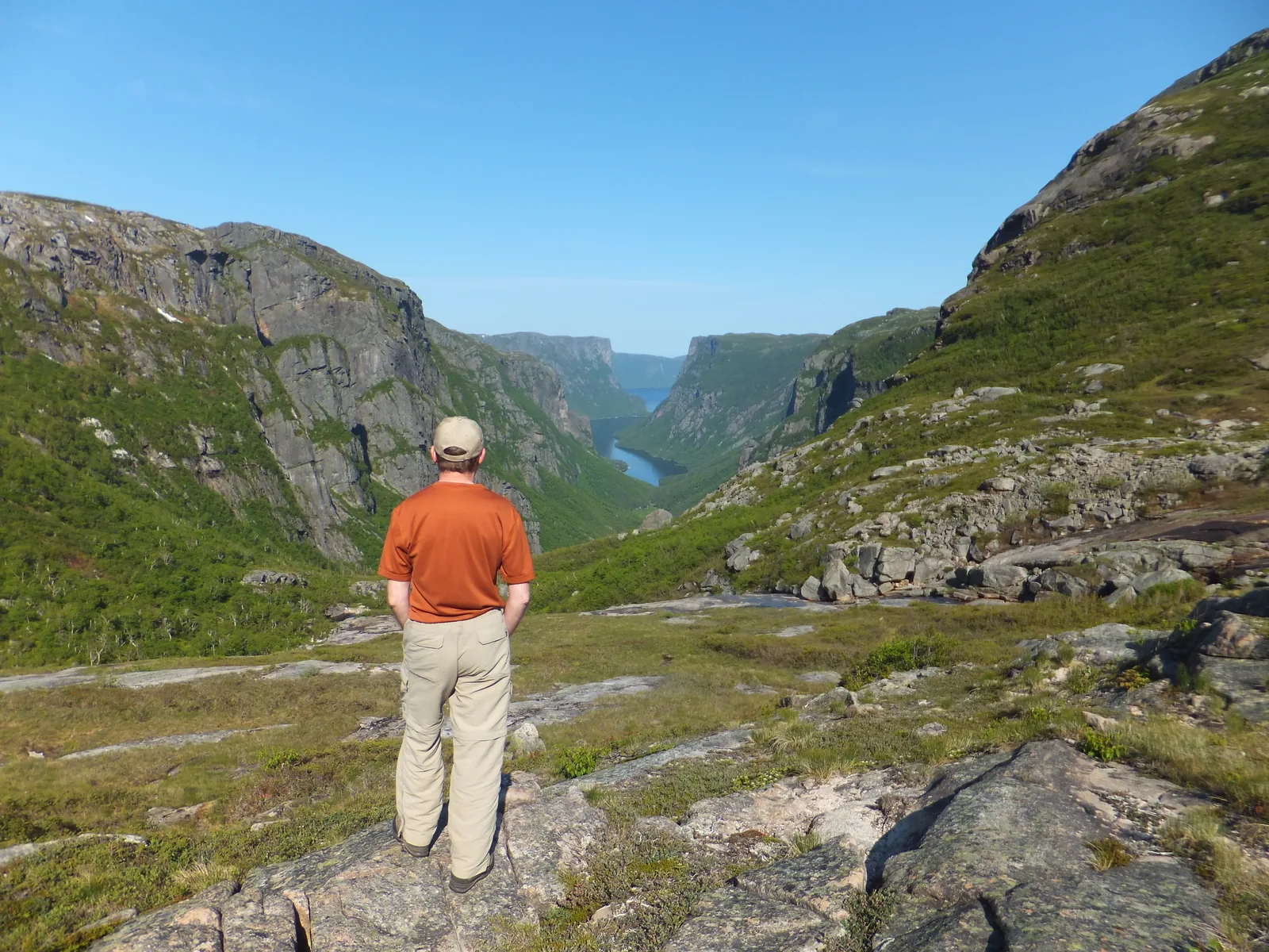 Ein Wanderer in oranger Jacke blickt vom felsigen Hang am Ende des Fjords hinab in das lange, schmale Tal des Western Brook Pond zwischen steilen Felswänden