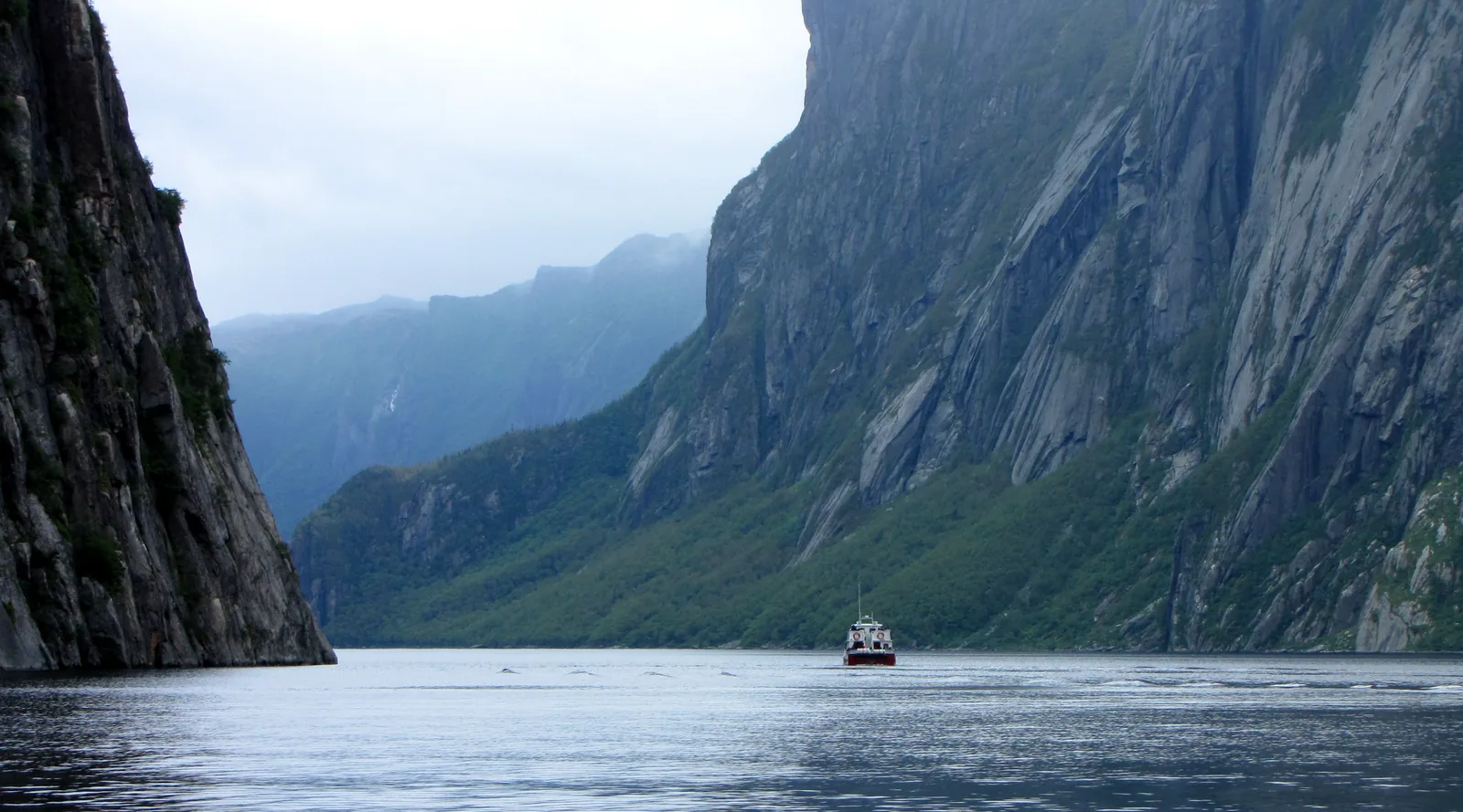 Senkrechte, dunkle Felswände rahmen den schmalen Western Brook Pond, in der Ferne fährt ein kleines Ausflugsboot auf dem stillen Wasser unter tiefen Wolken