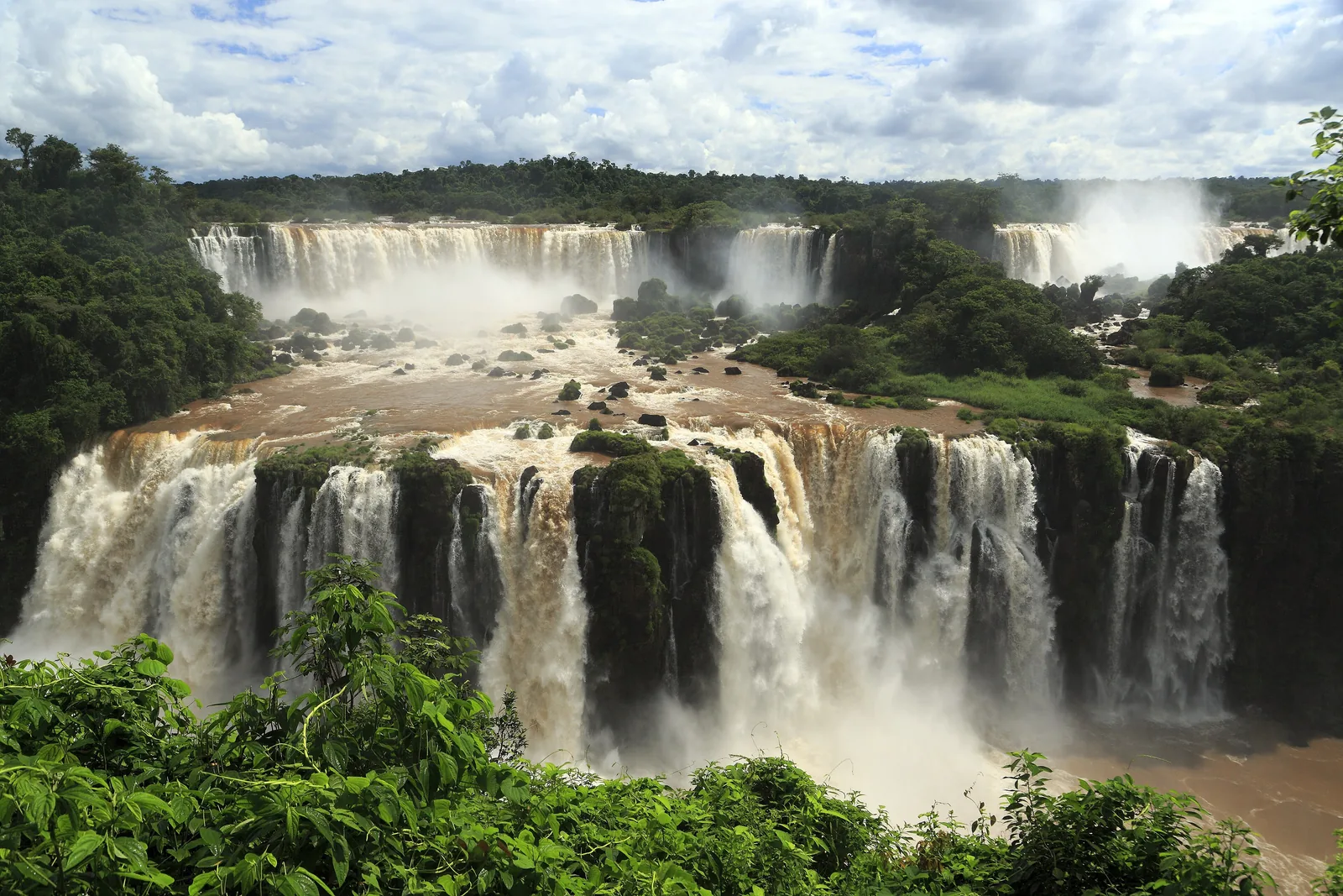 Von der brasilianischen Seite des Iguaçu-Nationalparks blickt man über eine Fallstufe aus vielen weißen Wasserfällen auf die Felsen und den Wald dahinter
