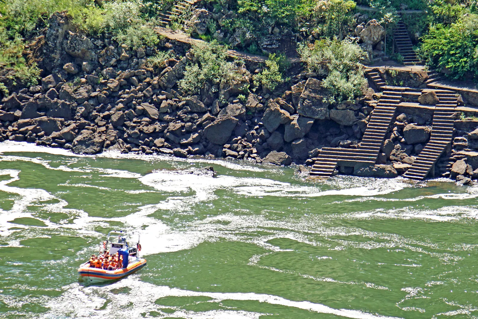 Ein Schlauchboot fährt auf grünem Wasser unterhalb der Iguazú-Fälle, am Ufer führt eine Treppe über Felsen nach oben