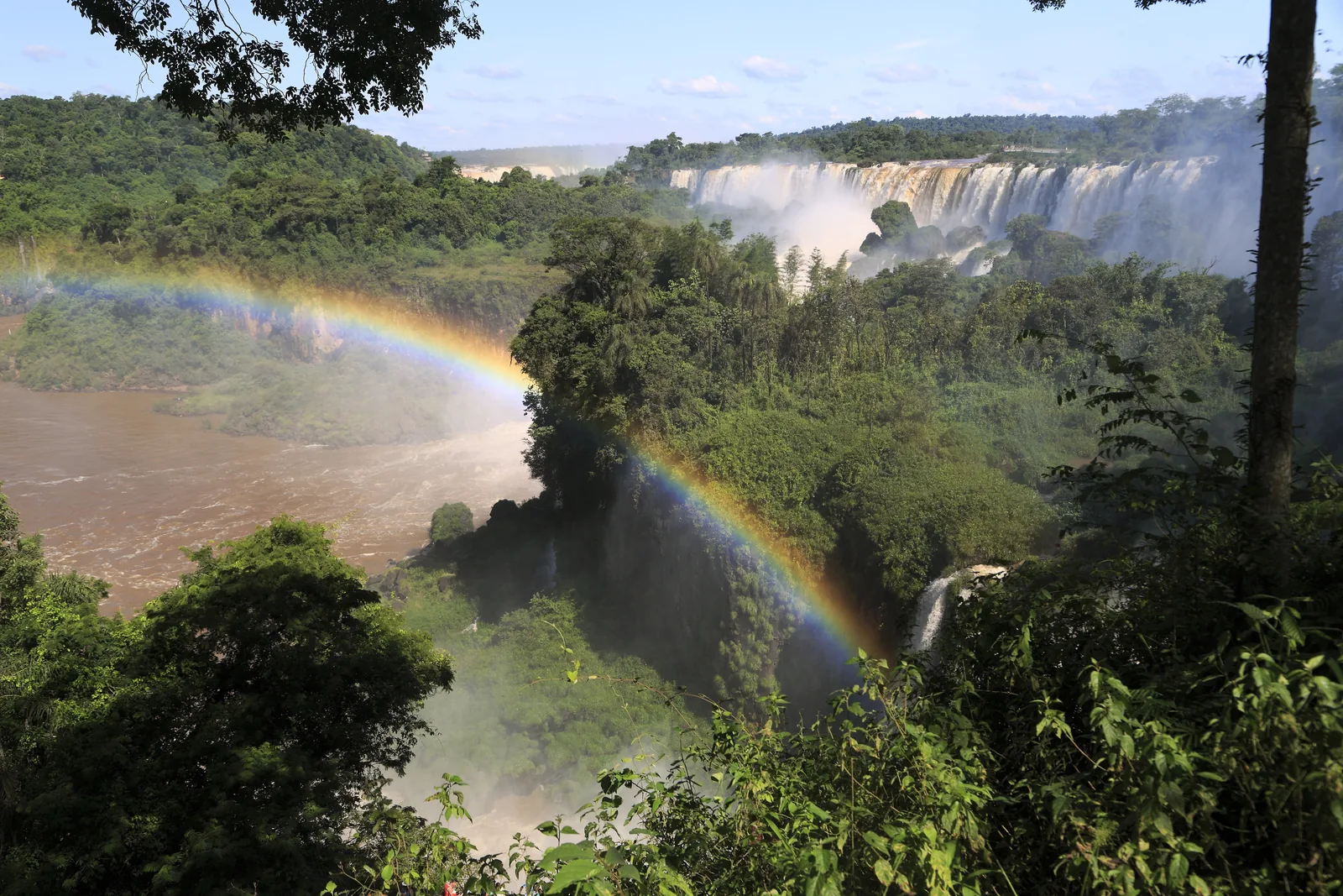 Blick vom oberen Rundweg im Iguazú-Nationalpark über bewaldete Felsen auf eine lange Reihe weißer Wasserfälle, davor ein Regenbogen über der Schlucht