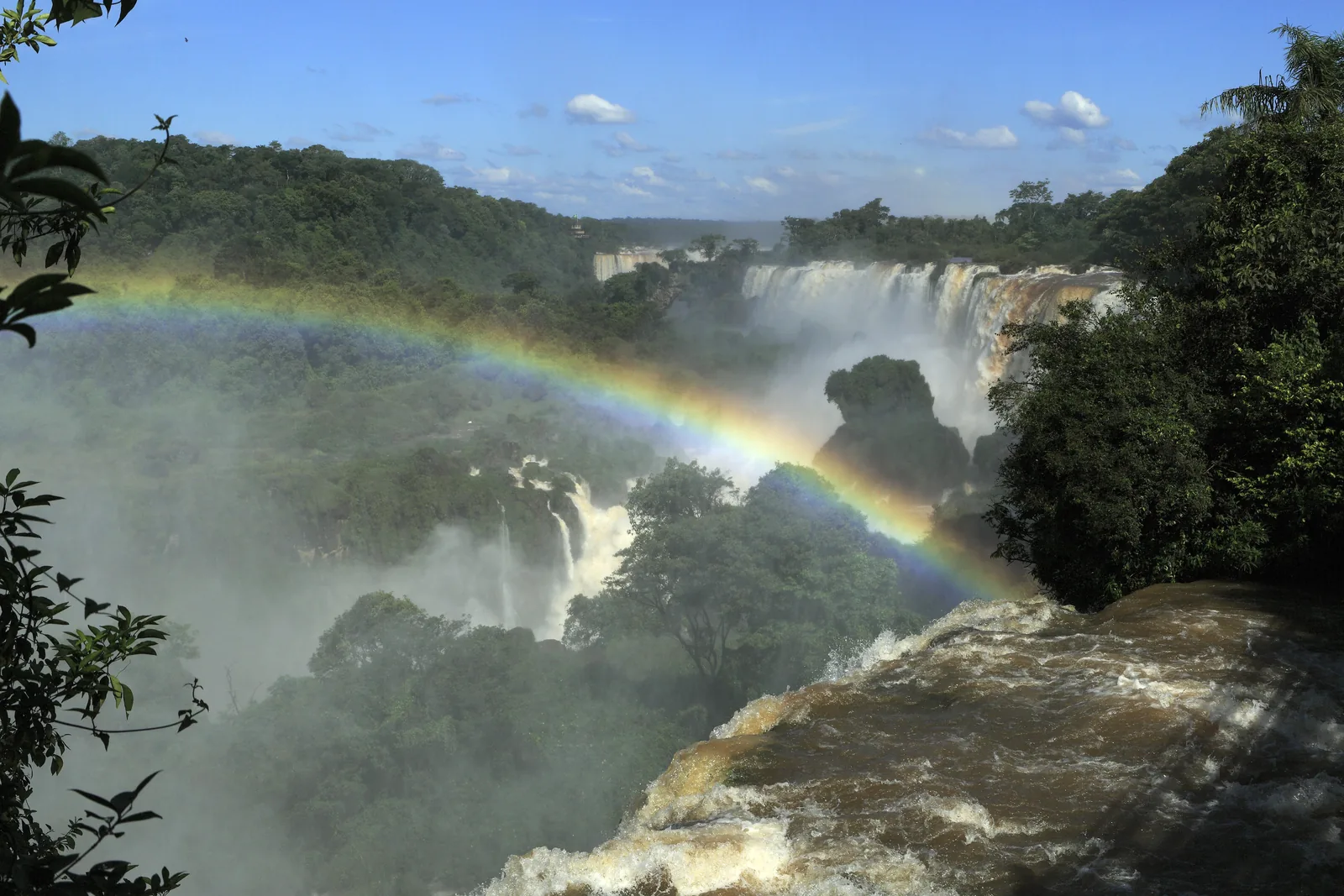 Vom oberen Rundweg der Iguazú-Wasserfälle spannt sich ein Regenbogen über die Schlucht, dahinter stürzen Fälle aus dem dichten Wald