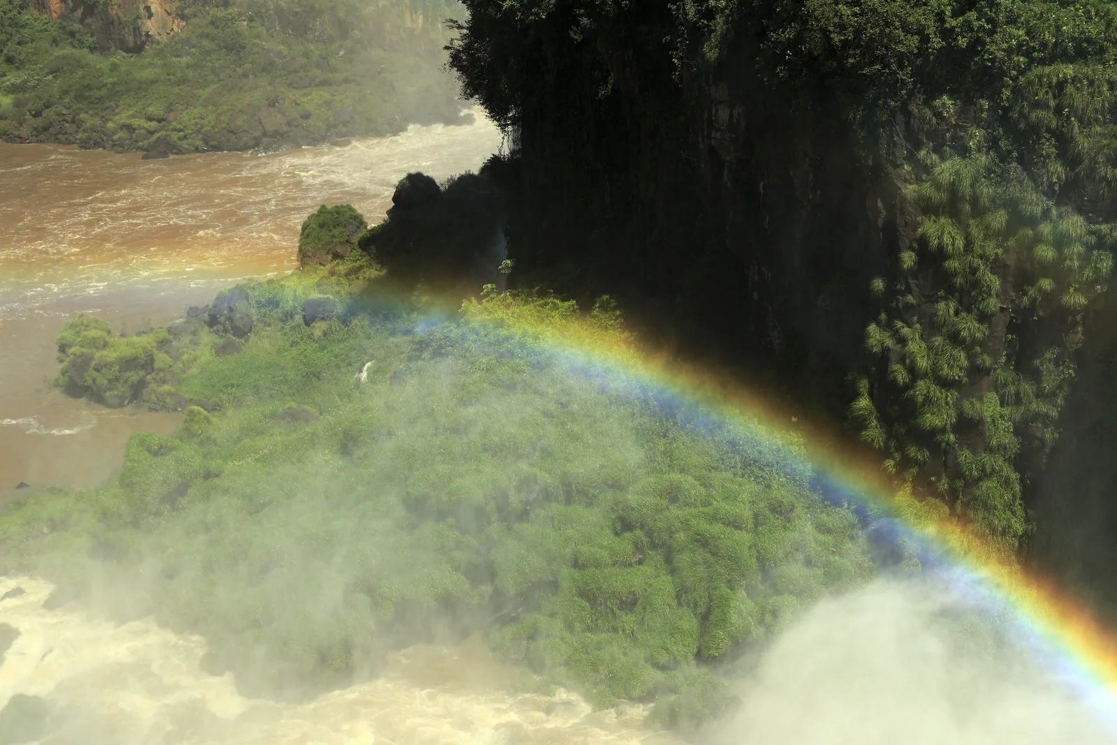 Ein kräftiger Regenbogen steht vor einer bewaldeten Felswand über der Gischt der Iguazú-Wasserfälle