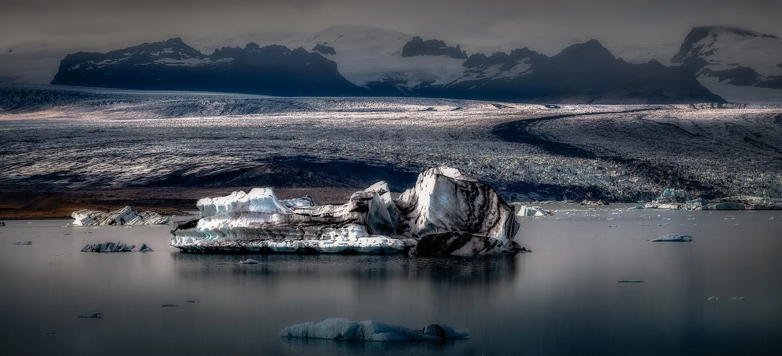 Ein dunkel gestreifter Eisberg treibt im Jökulsárlón vor der breiten Zunge des Breiðamerkurjökull