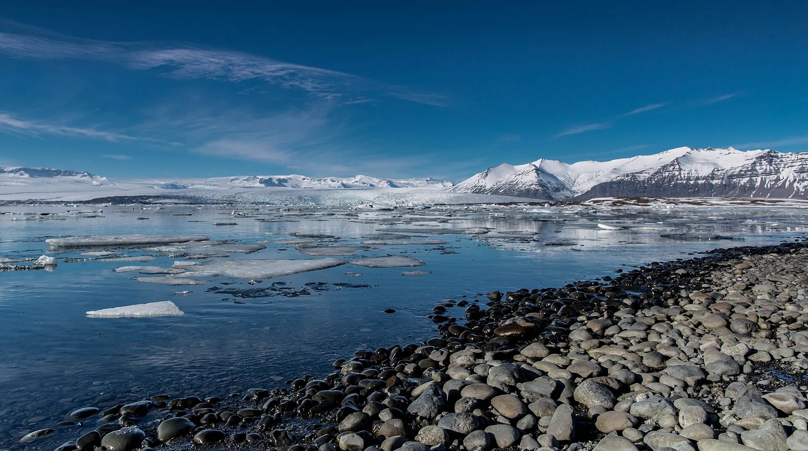 Die Gletscherlagune Jökulsárlón mit treibenden Eisbergen vor dem Breiðamerkurjökull, im Vordergrund ein Kiesufer, dahinter verschneite Berge
