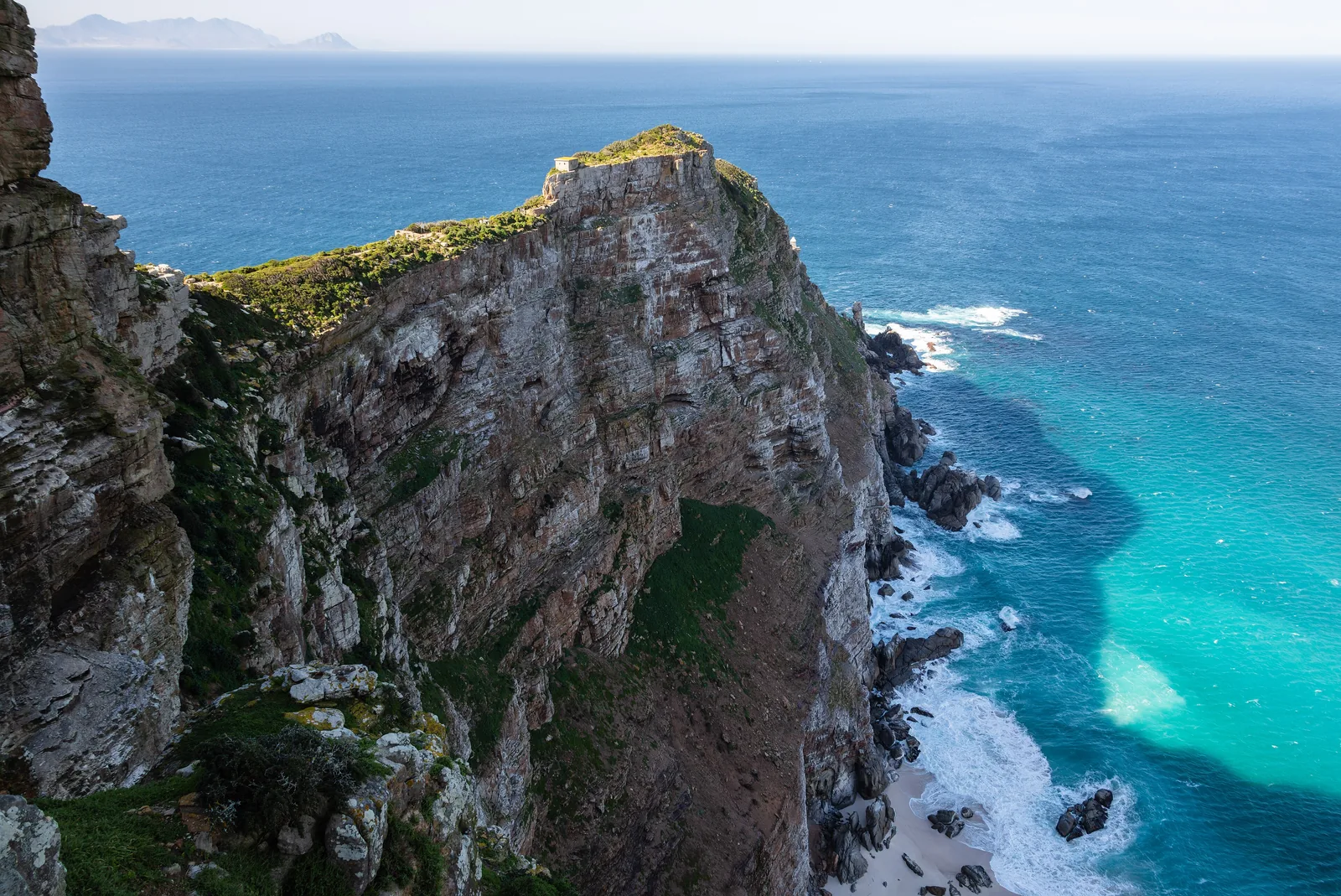 Steile, grün bewachsene Klippen am Kap der Guten Hoffnung fallen bei Cape Point in türkisfarbenes Meer mit weißer Brandung ab