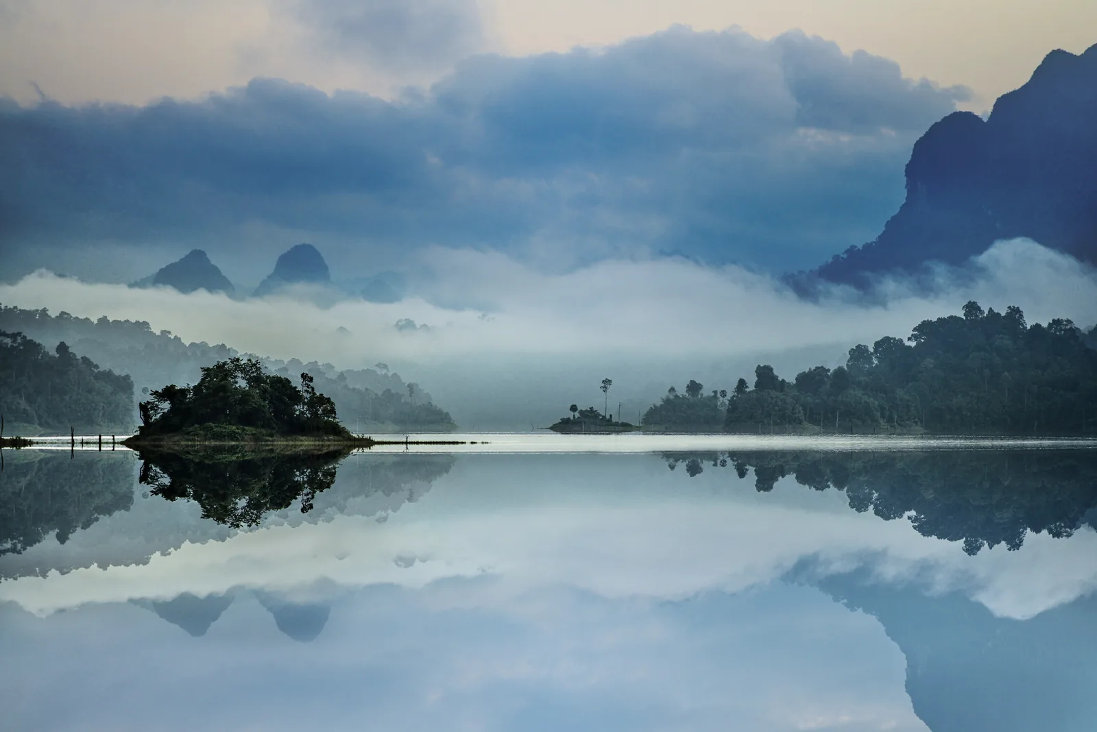 Spiegelglatter See im Morgennebel, eine kleine bewaldete Insel und dahinter die Silhouetten zackiger Kalkberge spiegeln sich im Wasser