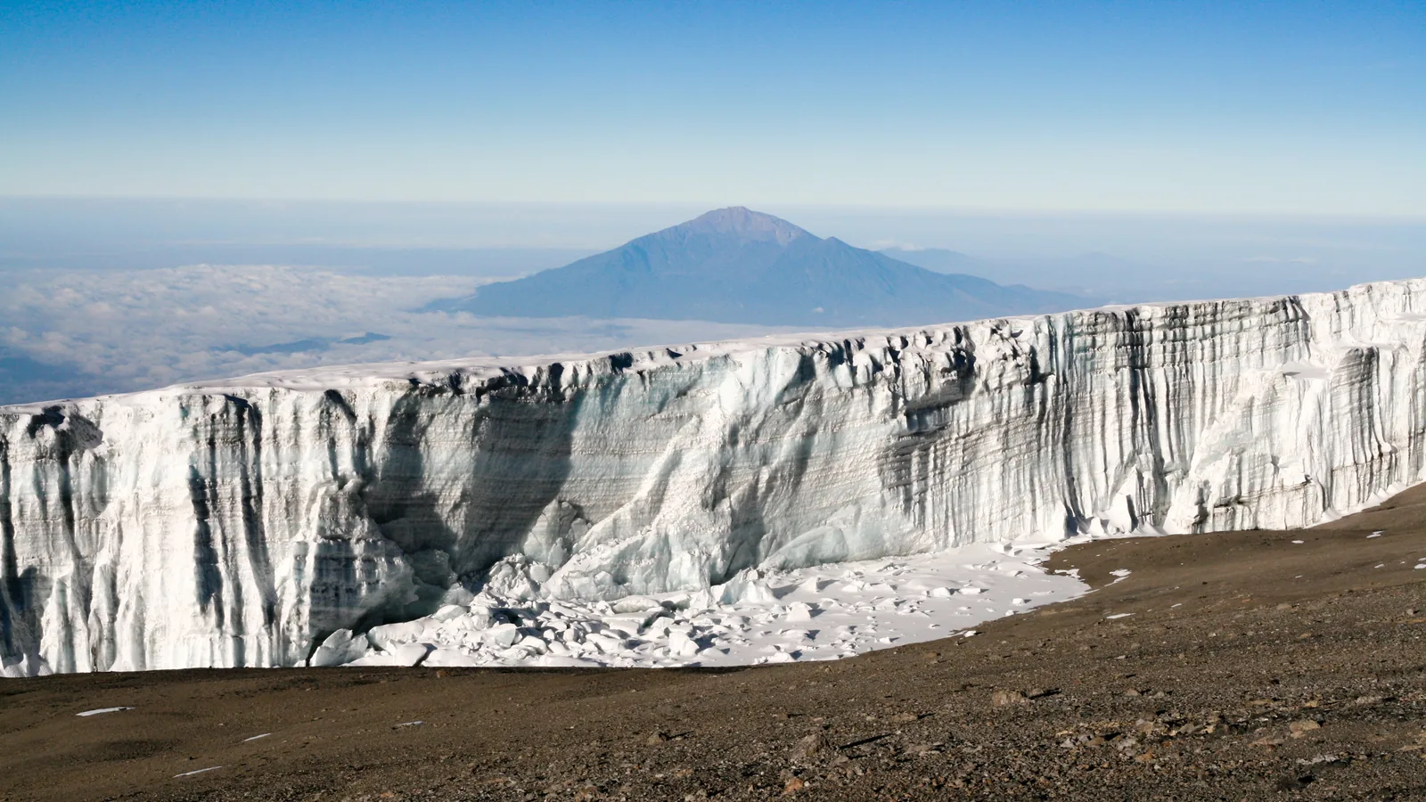 Eine senkrechte Gletscherwand nahe dem Uhuru Peak im Kilimandscharo-Nationalpark, in der Ferne der Mount Meru über den Wolken