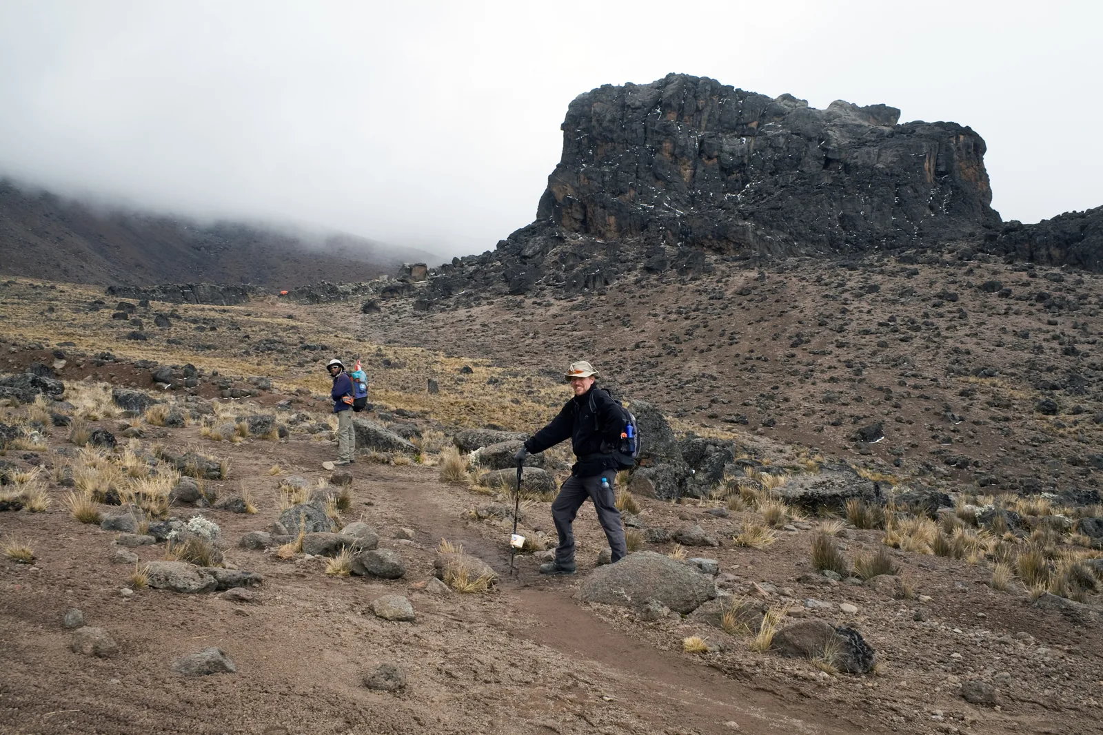 Wanderer mit Stöcken vor dem Lava Tower im Kilimandscharo-Nationalpark, einem dunklen Felsturm über Geröll und Wolken