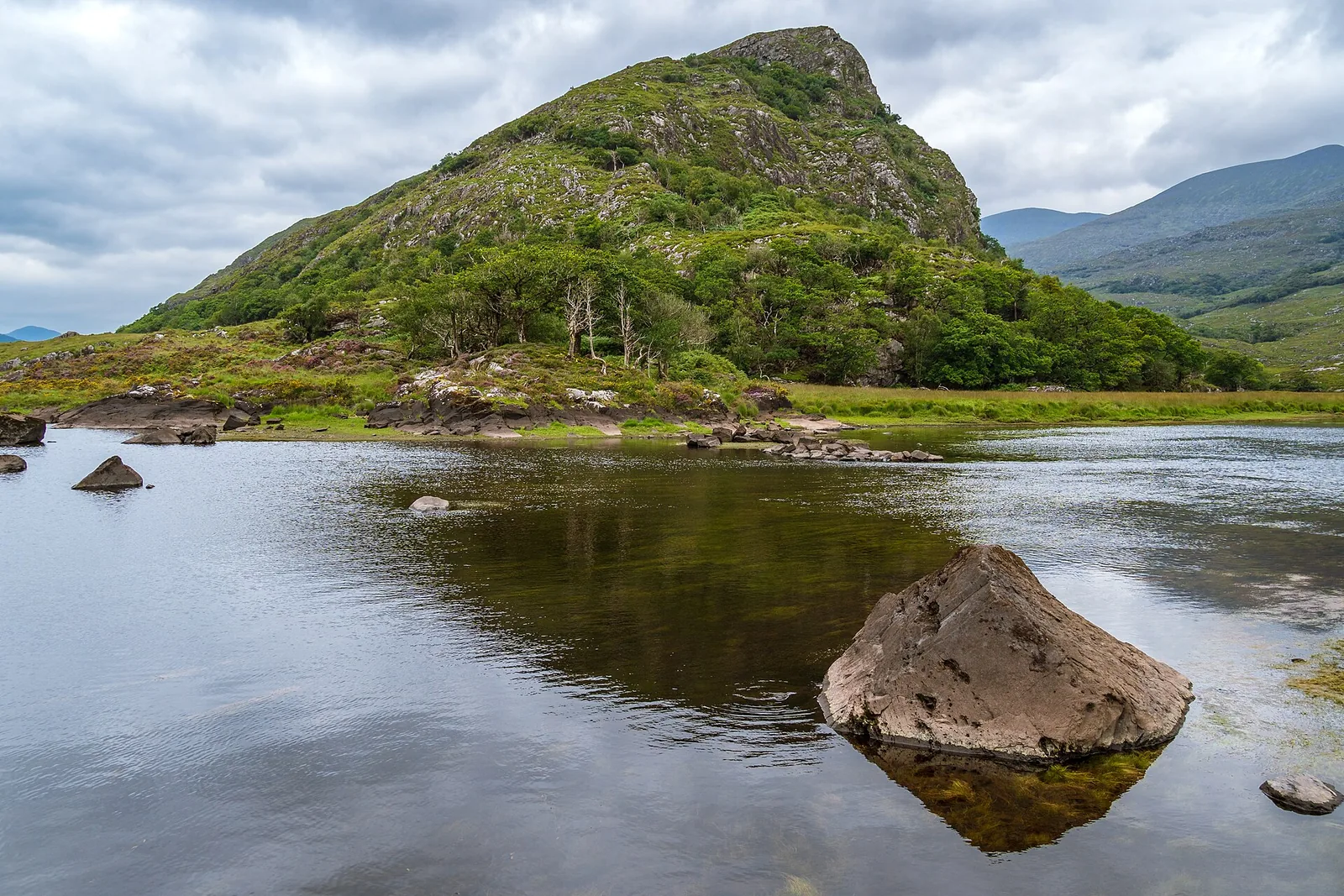 Ein See im Killarney-Nationalpark zwischen grünen Hügeln und den Bergen von Kerry unter einem wolkigen Himmel