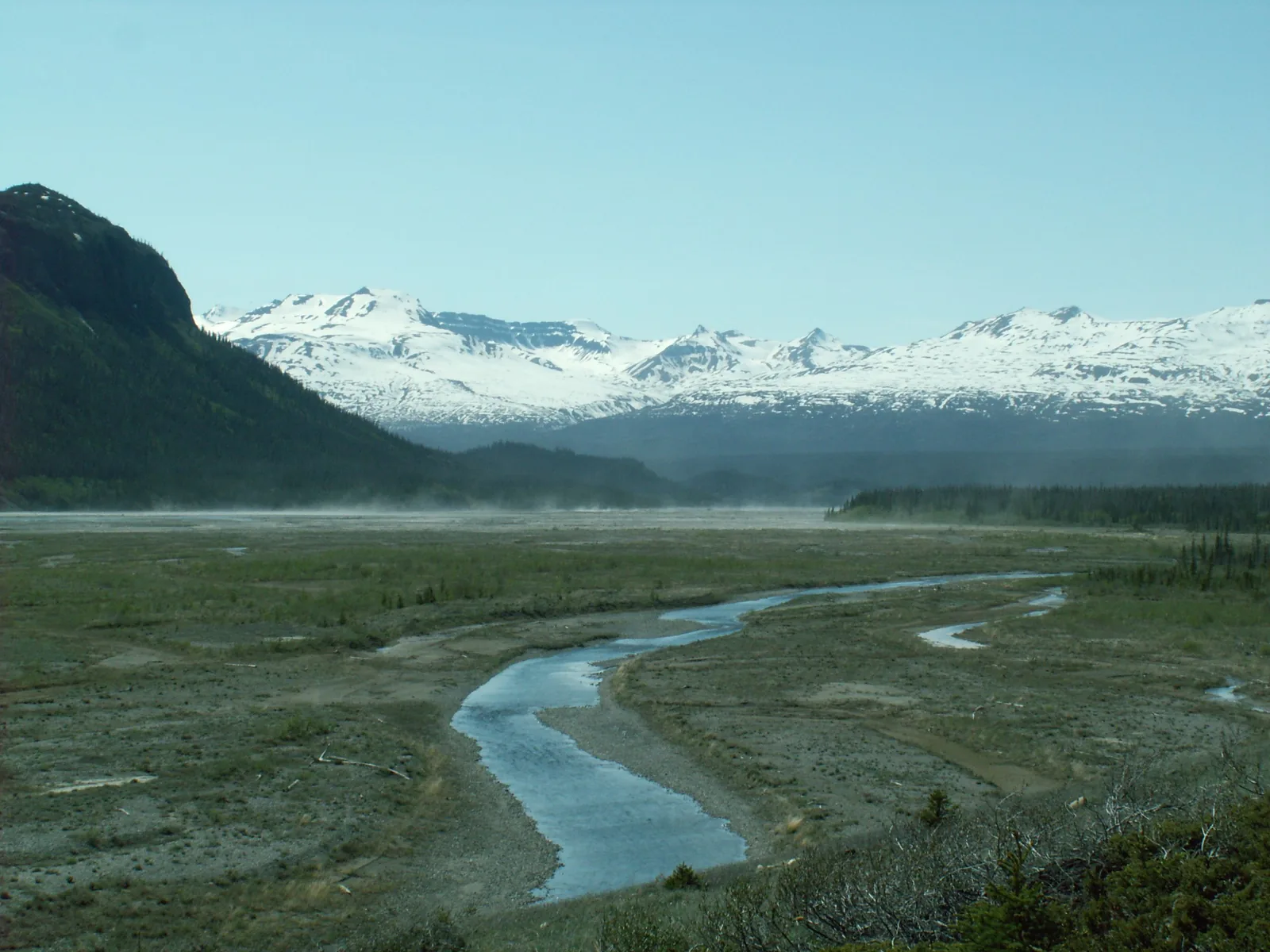 Der verzweigte Alsek River in einer weiten Schotterebene, dahinter eine schneebedeckte Bergkette im Dunst