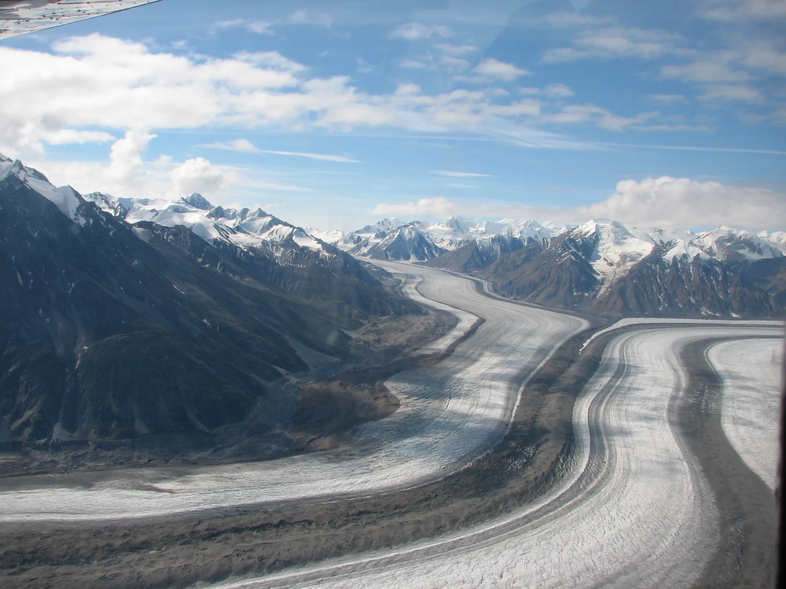 Der Kaskawulsh-Gletscher zieht mit dunklen Mittelmoränen durch ein breites Tal, ringsum schneebedeckte Gipfel
