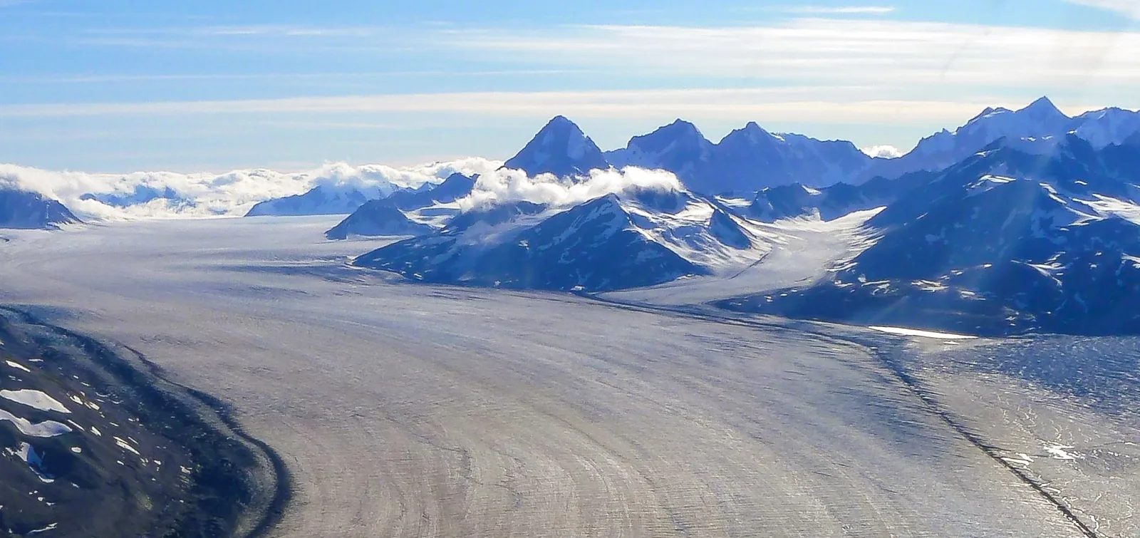 Der Lowell-Gletscher zieht als breiter Eisstrom durch das Bild, dahinter der Ulu Mountain und weitere Gipfel über einer Wolkenschicht