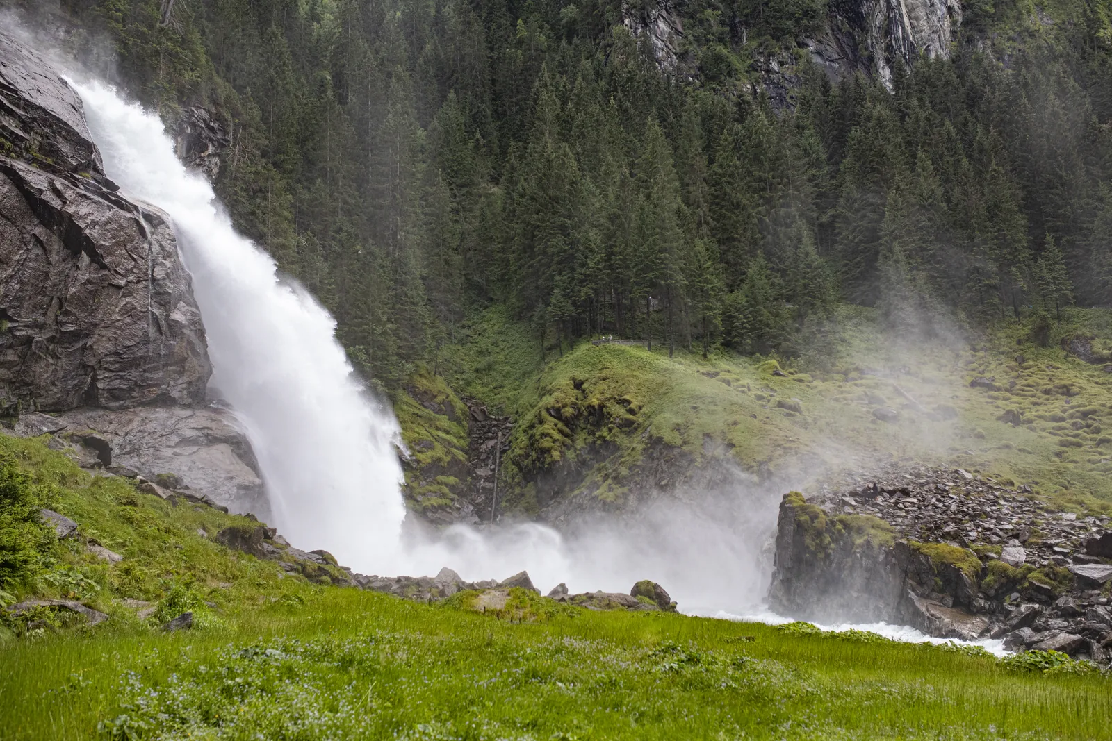 Der untere Krimmler Wasserfall stürzt über eine Felsstufe, Gischt zieht über die Wiese am Fuß der Fälle im Nationalpark Hohe Tauern