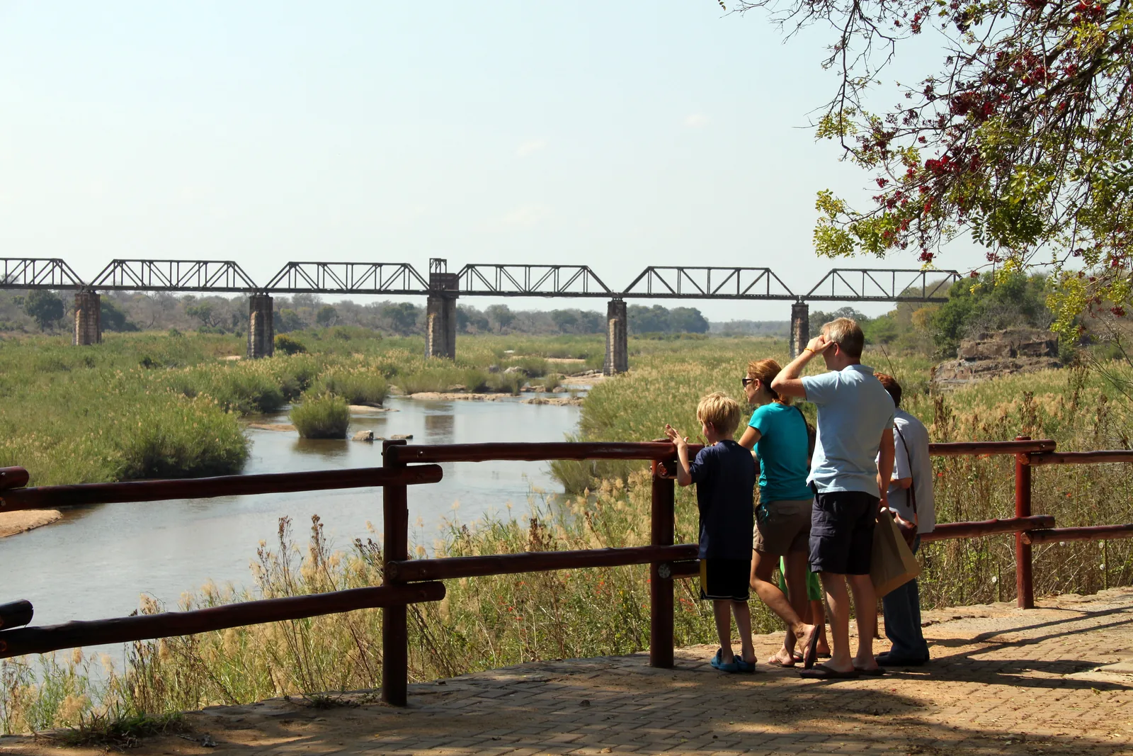 Besucher stehen an einem Holzgeländer über dem Sabie River, im Hintergrund eine alte Eisenbahnbrücke aus Stahlfachwerk