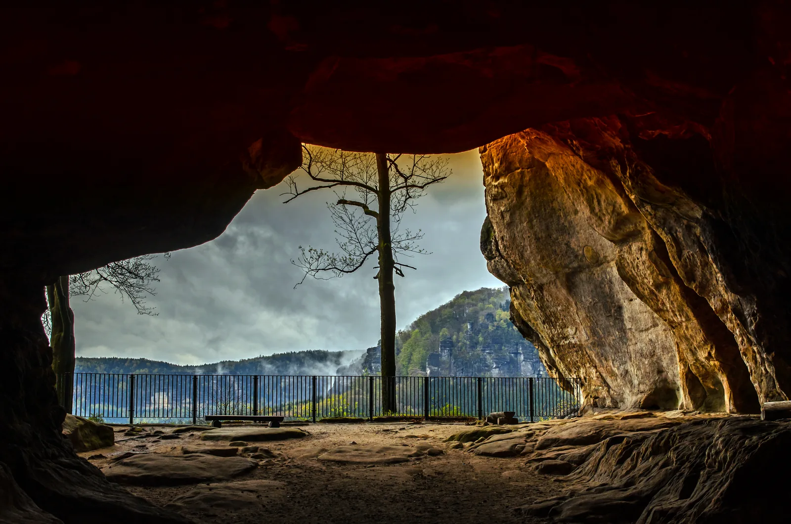 Blick durch das Felsentor Kuhstall im Nationalpark Sächsische Schweiz auf die bewaldeten Affensteine, im Vordergrund das Geländer der Aussicht