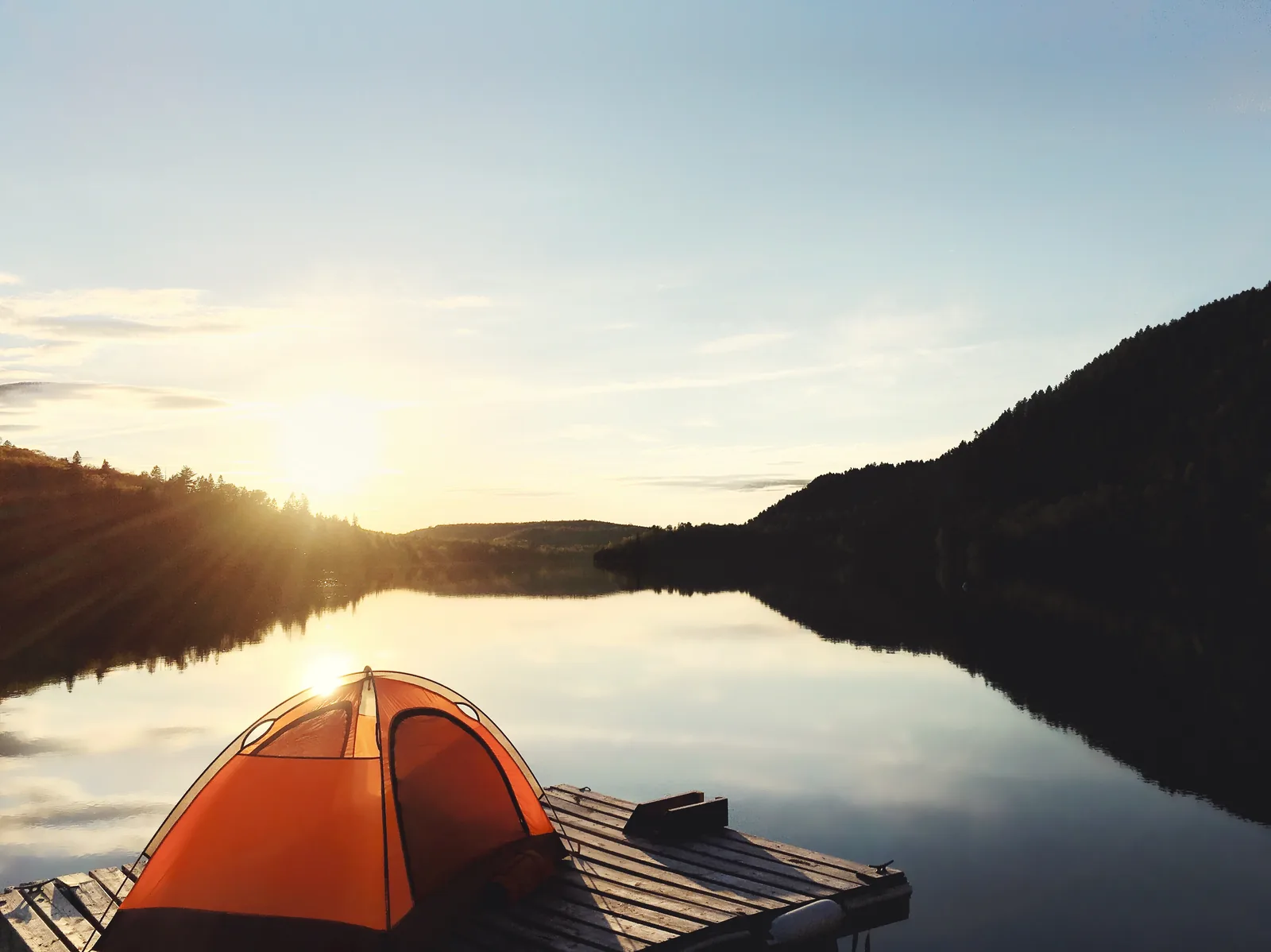 Ein orangefarbenes Zelt steht auf einer Holzplattform am Seeufer, die Sonne geht hinter den bewaldeten Hügeln auf und spiegelt sich im stillen Wasser